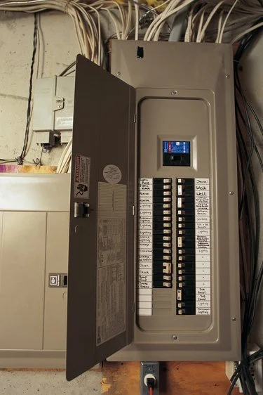 Electrical breaker panel with open door showing circuit breakers and a digital display, surrounded by various cables in an electrical utility room.