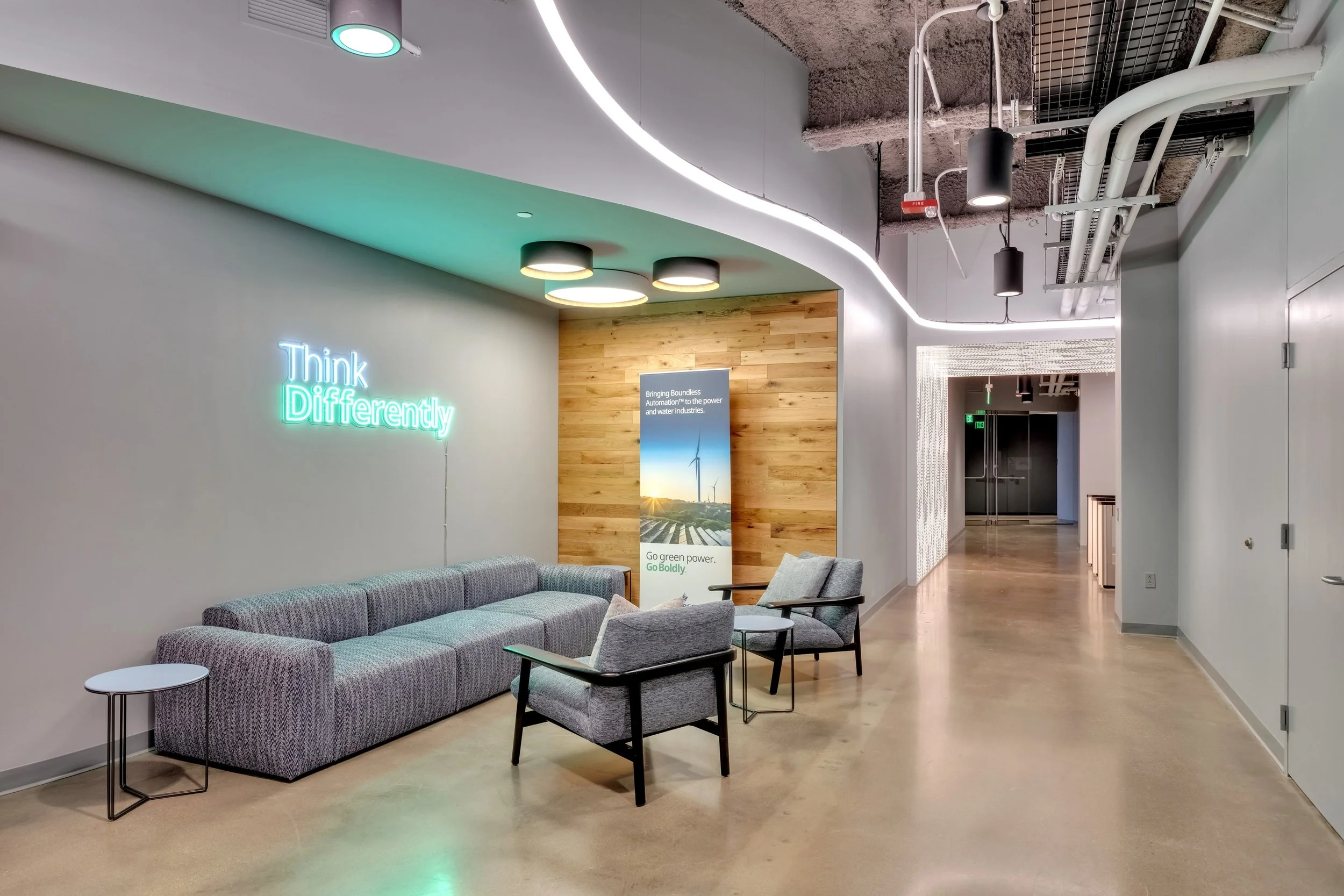 Modern office lobby with a neon sign reading "Think Differently" on a wall, a gray couch, two armchairs, a small side table, a wooden accent wall with a poster about green power, and suspended ceiling lights.