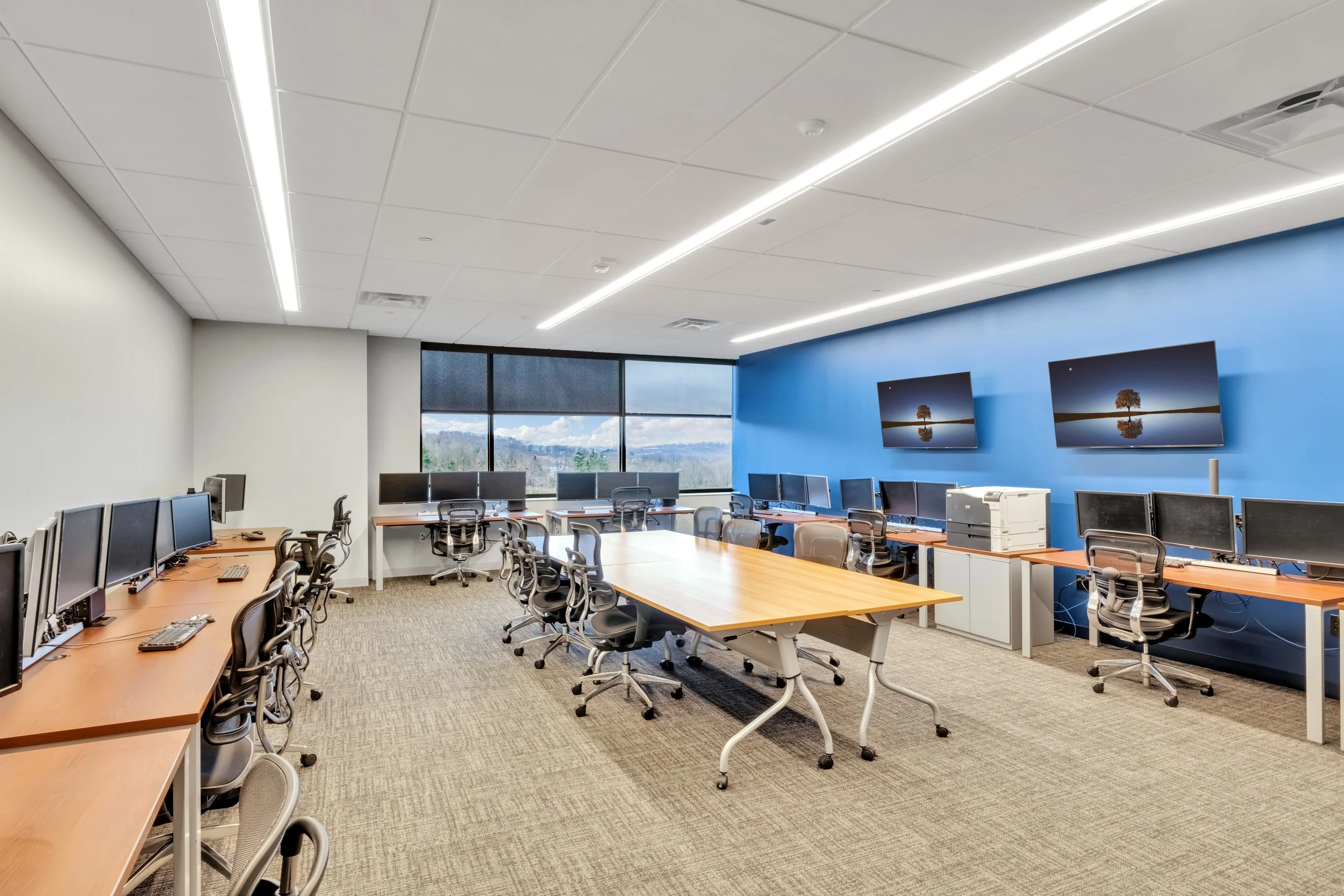 Empty computer lab with multiple desktop computers, black office chairs, a wooden table, large windows showing scenic mountain view, and two wall-mounted monitors on a blue wall.