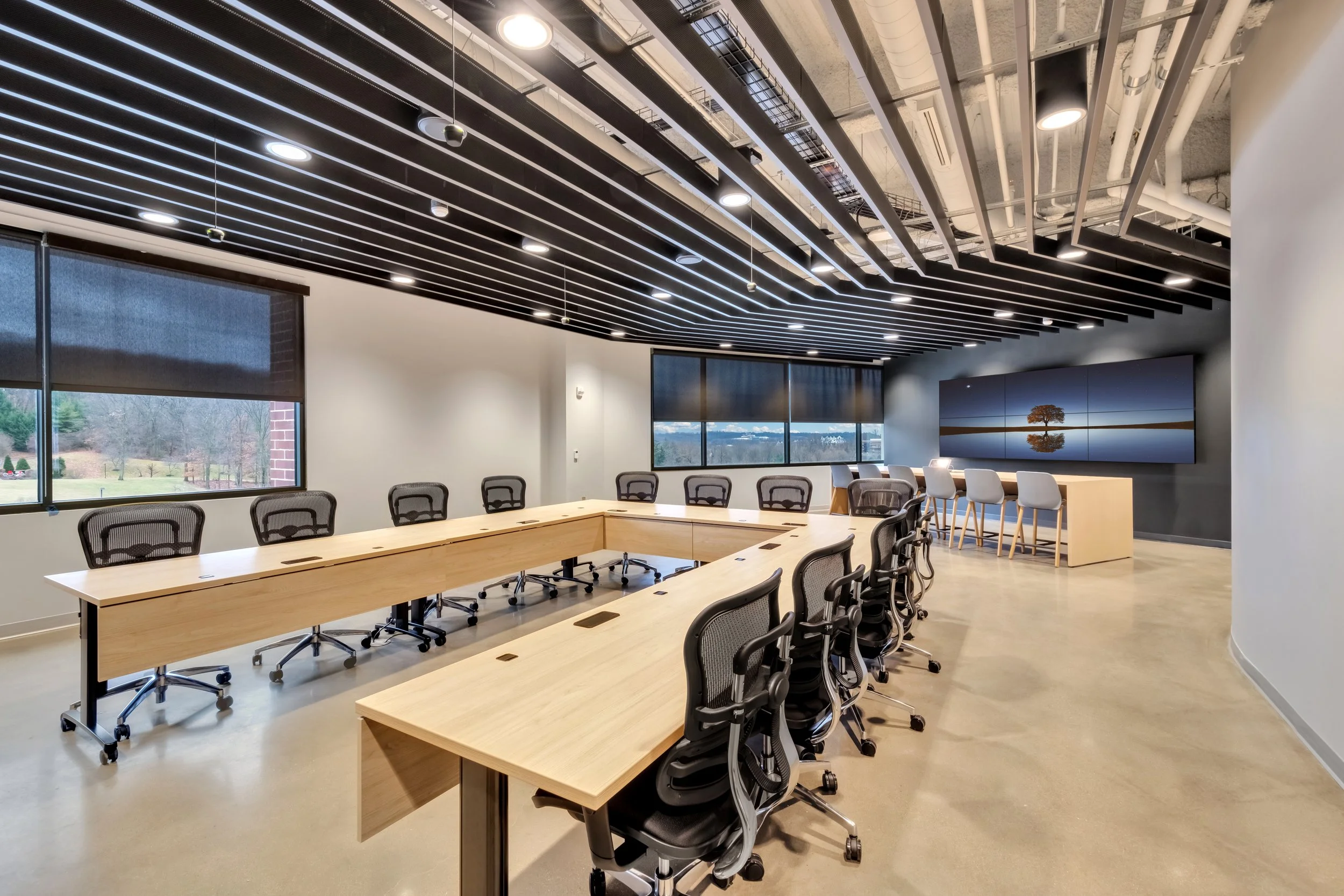 Modern conference room with U-shaped wooden table, black office chairs, large windows, and a multi-panel display showing a tree with water reflection