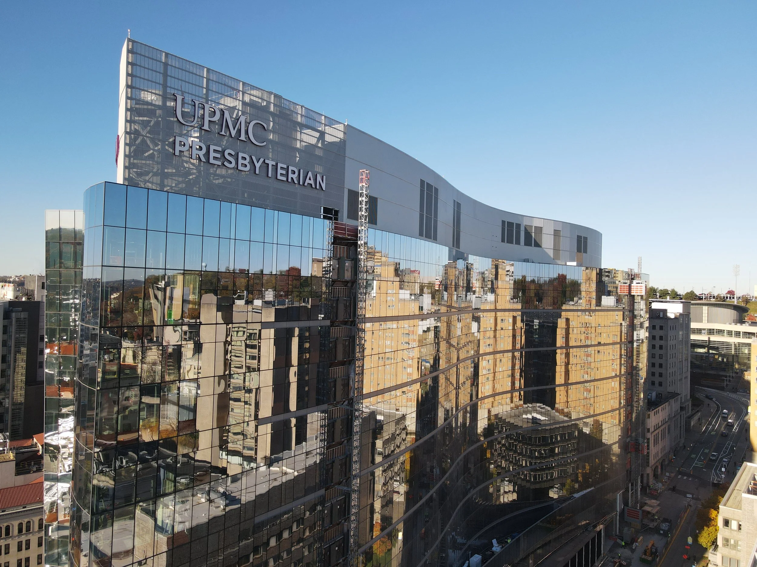 Reflected glass skyscraper with the sign 'UPMC PRESBYTERIAN' on top, showing distorted views of surrounding buildings and city streets below in bright daylight.