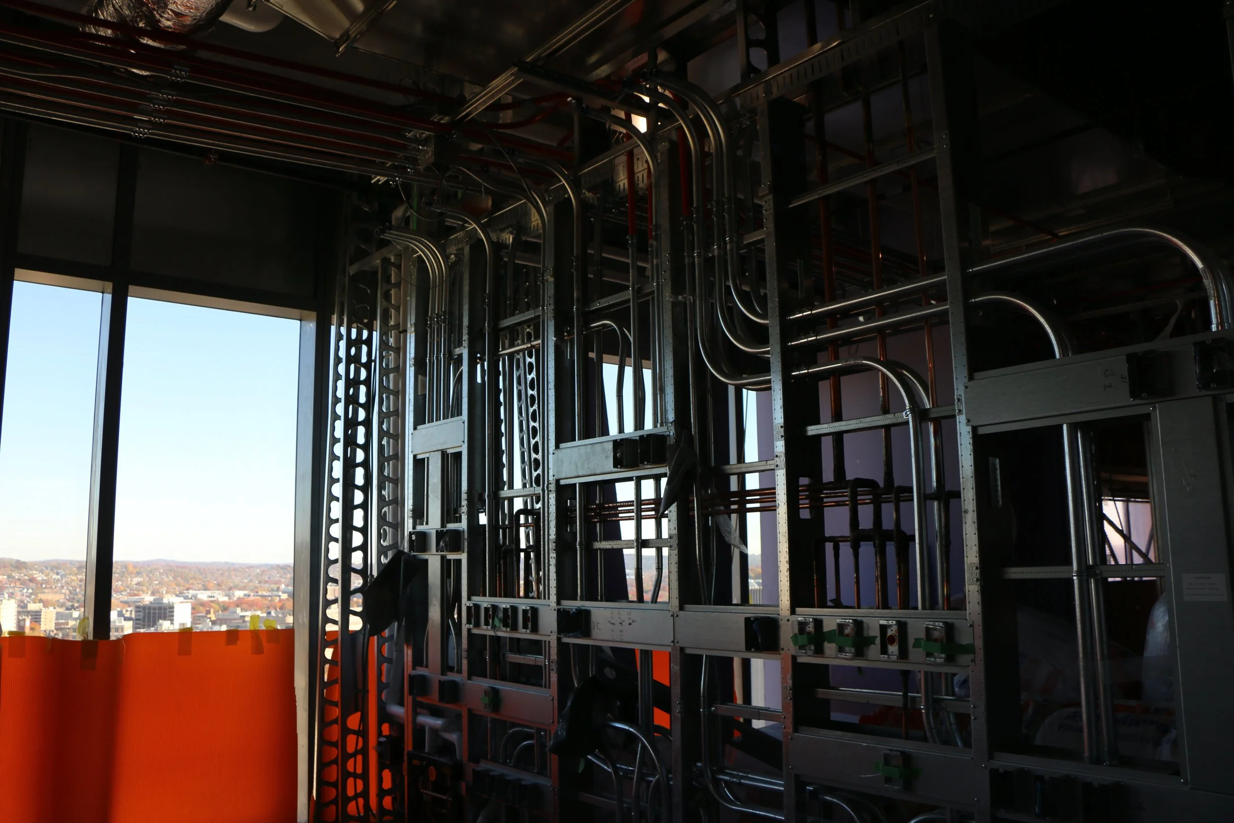 Interior of a building under construction with visible metal framing, wiring, and open window showing cityscape outside.