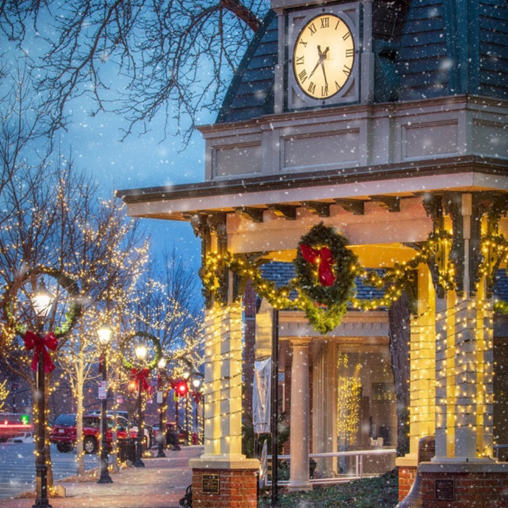 A decorated town square during winter with Christmas lights, wreaths, and snow falling, featuring a clock tower at dusk.