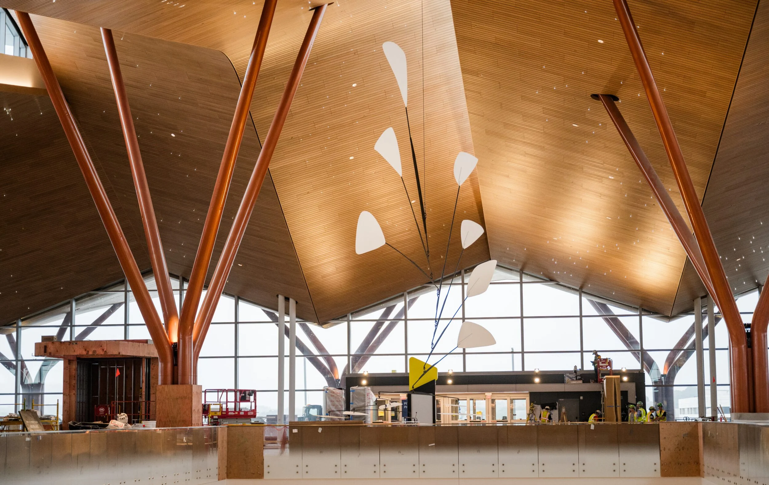 Interior view of a modern building under construction with a high wooden ceiling, large glass windows, orange structural supports, and a contemporary white art sculpture hanging from the ceiling. Several construction workers are present in the background.
