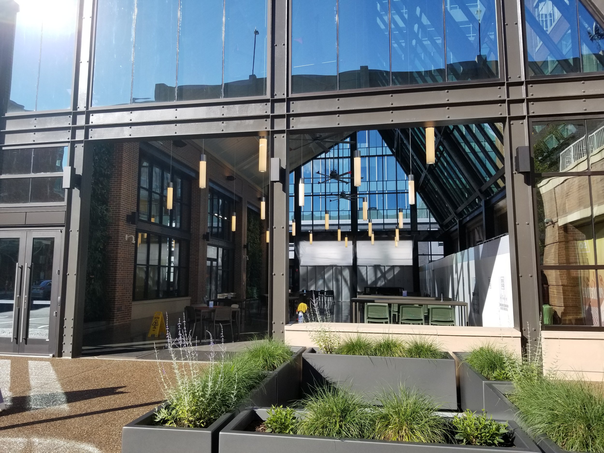 Exterior view of a modern glass building with an interior space visible, featuring hanging lights, tables, and chairs, with plants and planters outside.