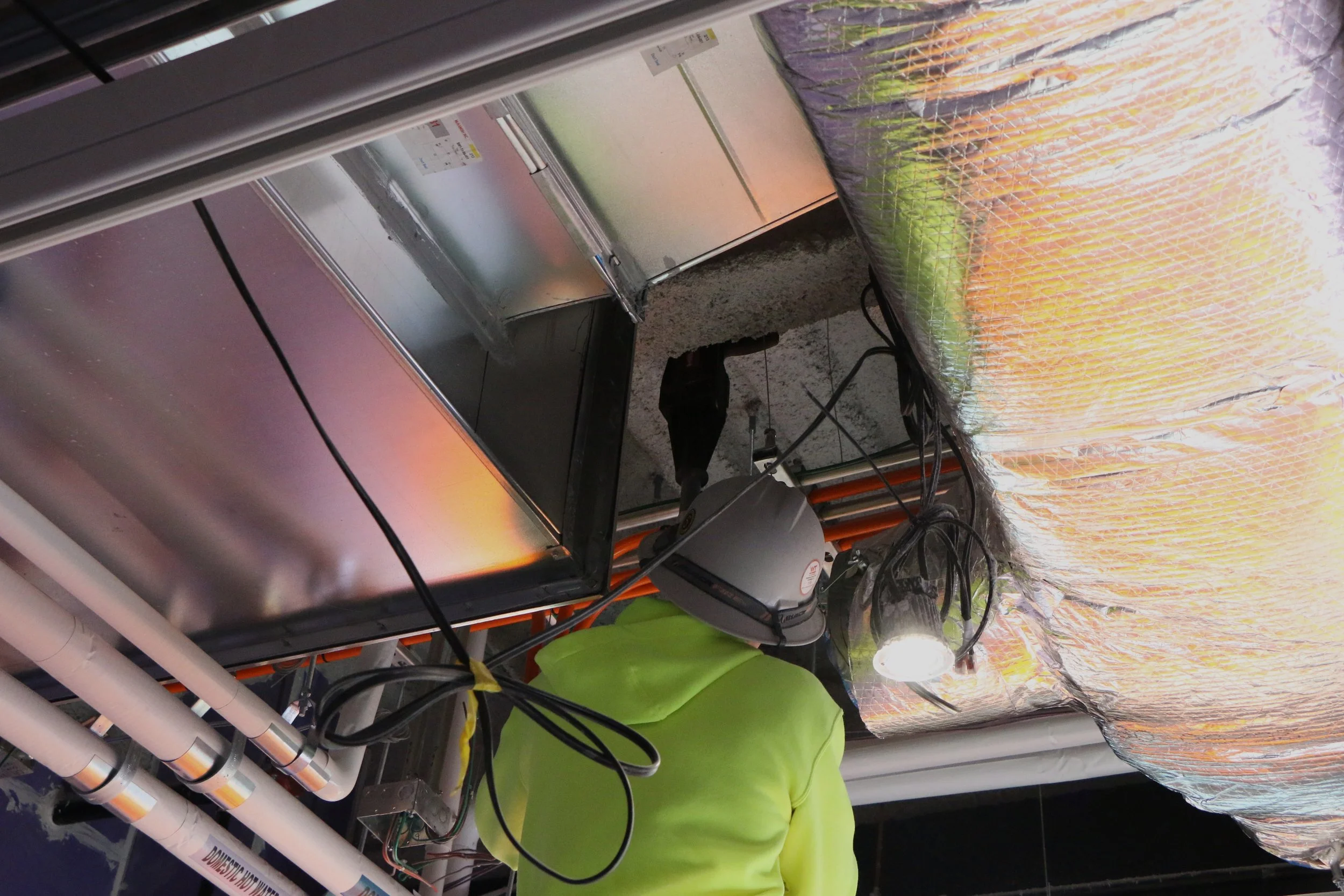 Worker in neon green shirt installing or repairing equipment in a ceiling with bright lights and insulation material.