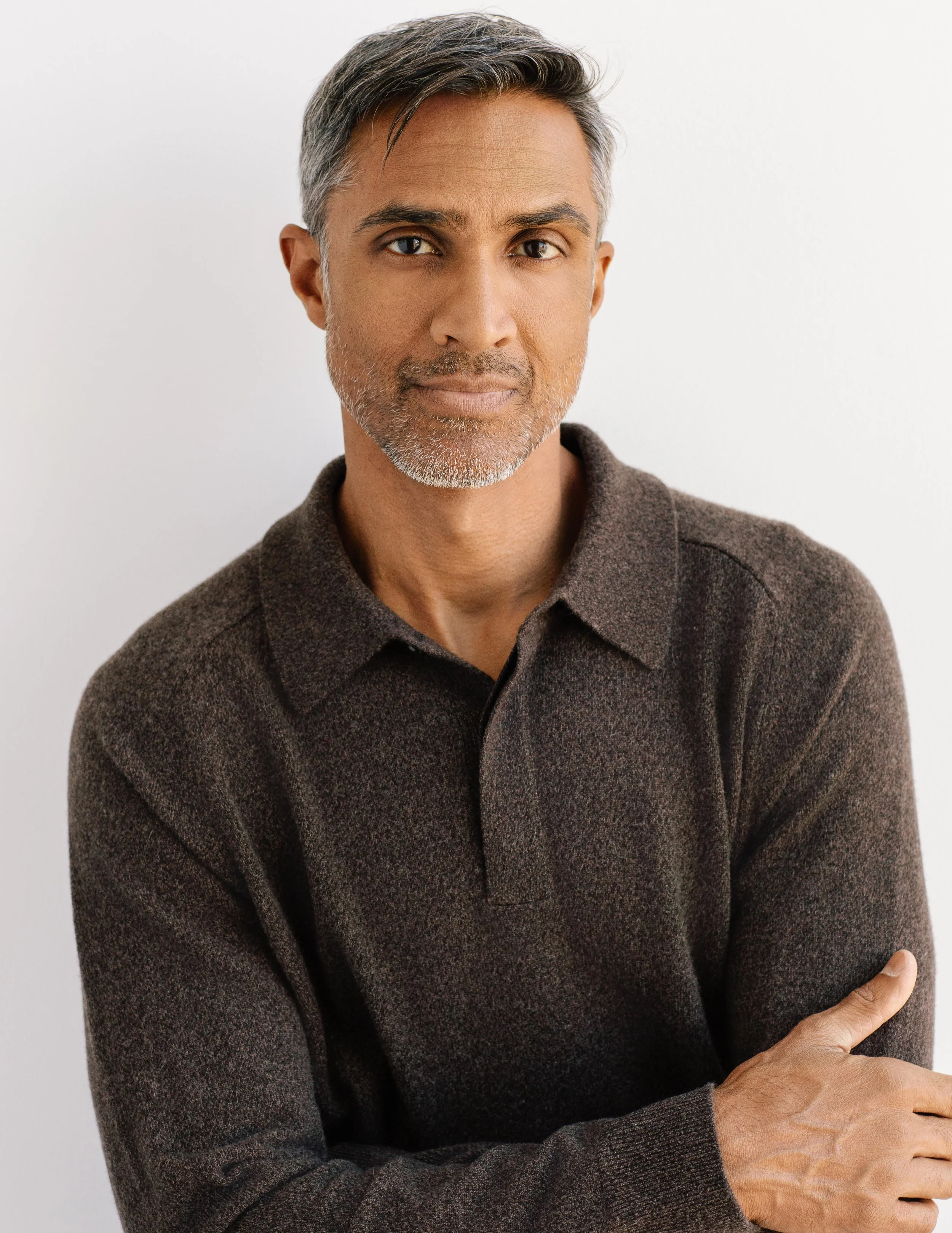 Dylan Jawahir, licensed acupuncturist and massage therapist specializing in natural pain management wearing brown long sleeve polo and leaning against a wall with arms folded in front of him.