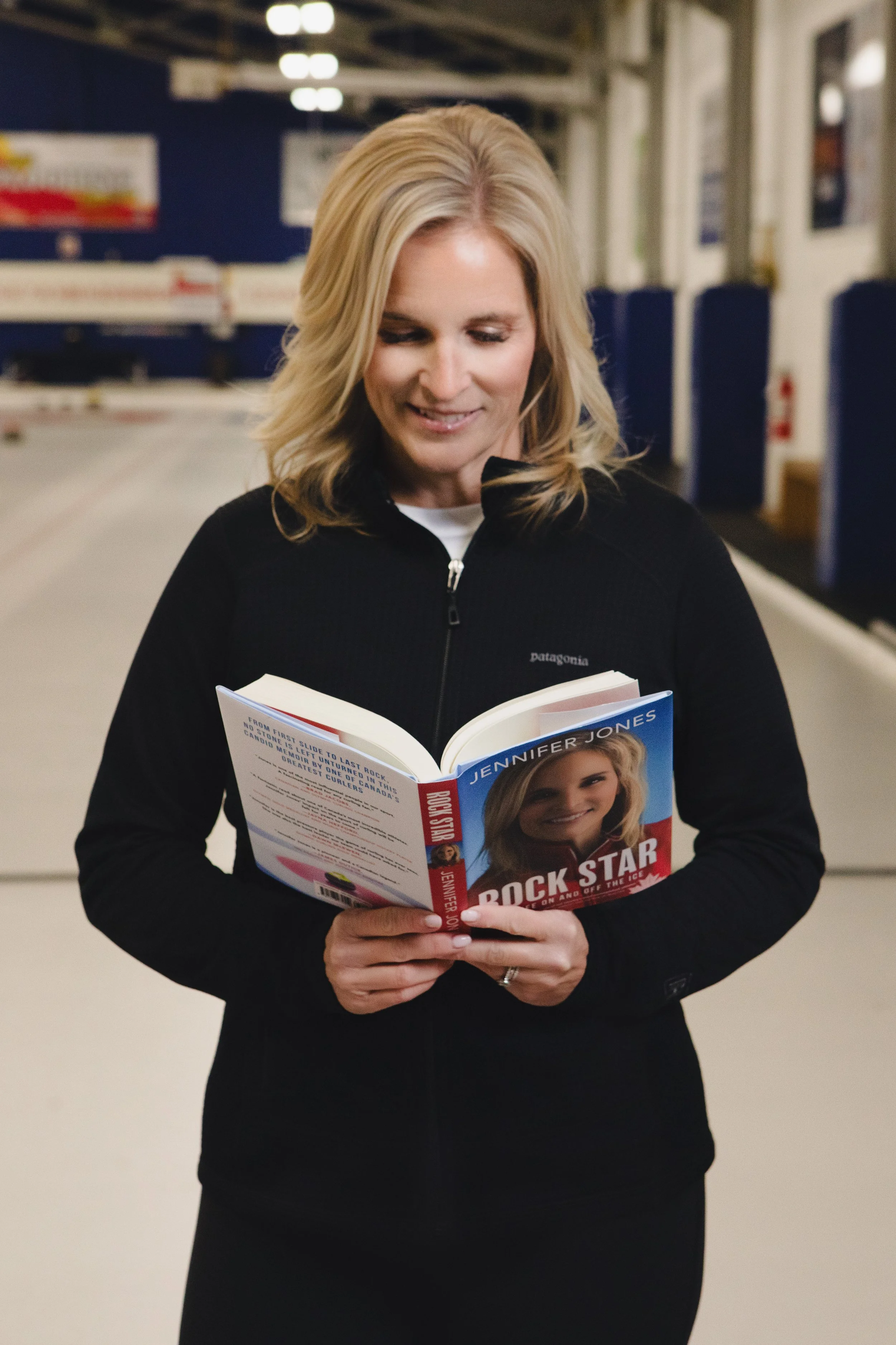 A blonde woman in a black jacket reads a book titled 'Rock Star' by Jennifer Jones on a train platform.
