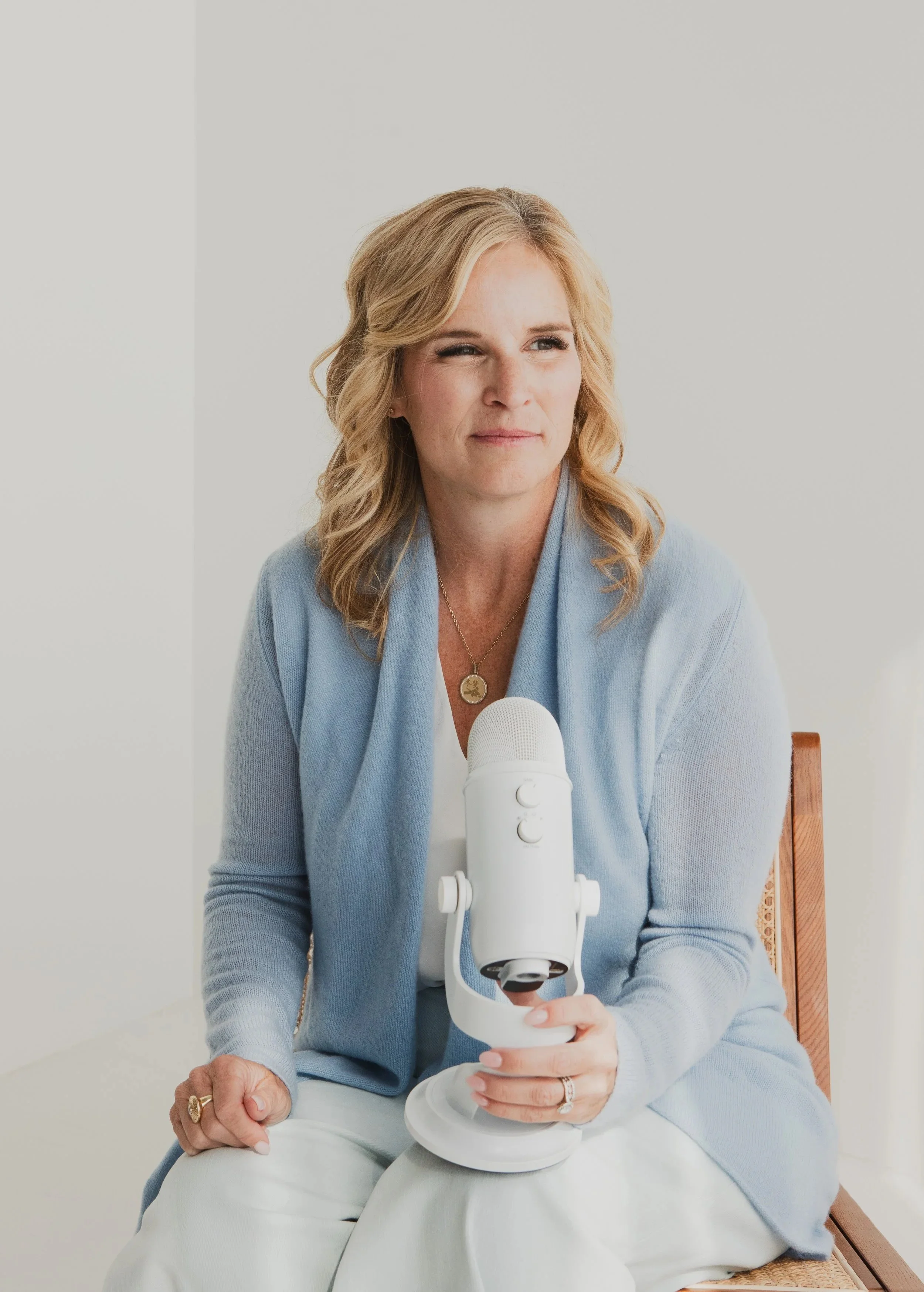 A woman with blonde hair sitting on a chair holding a white microphone, wearing a light blue cardigan and white pants, against a plain light background.