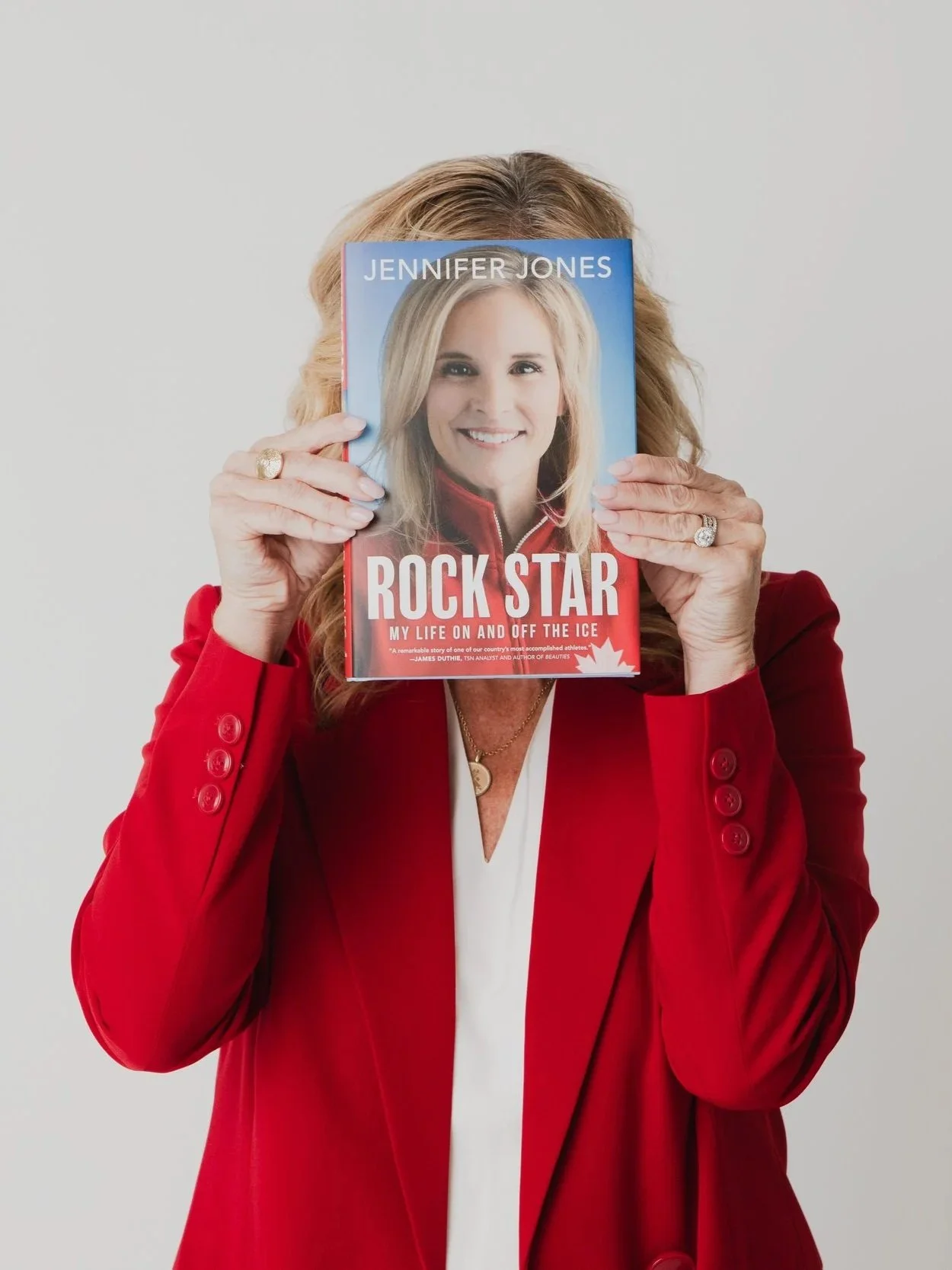 Woman in red blazer holding a book in front of her face, cover shows a smiling blonde woman, titled 'Jennifer Jones Rock Star', with a subtitle about her life.