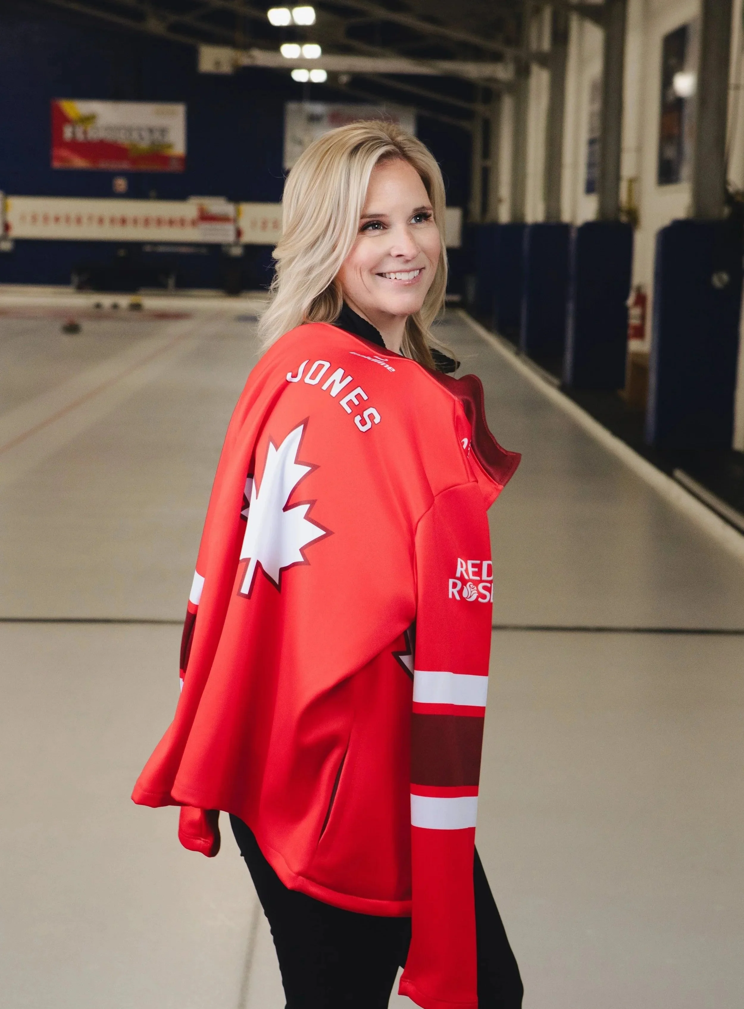 A smiling woman with blonde hair wearing a red hockey jersey with a maple leaf logo, standing in an indoor ice rink.