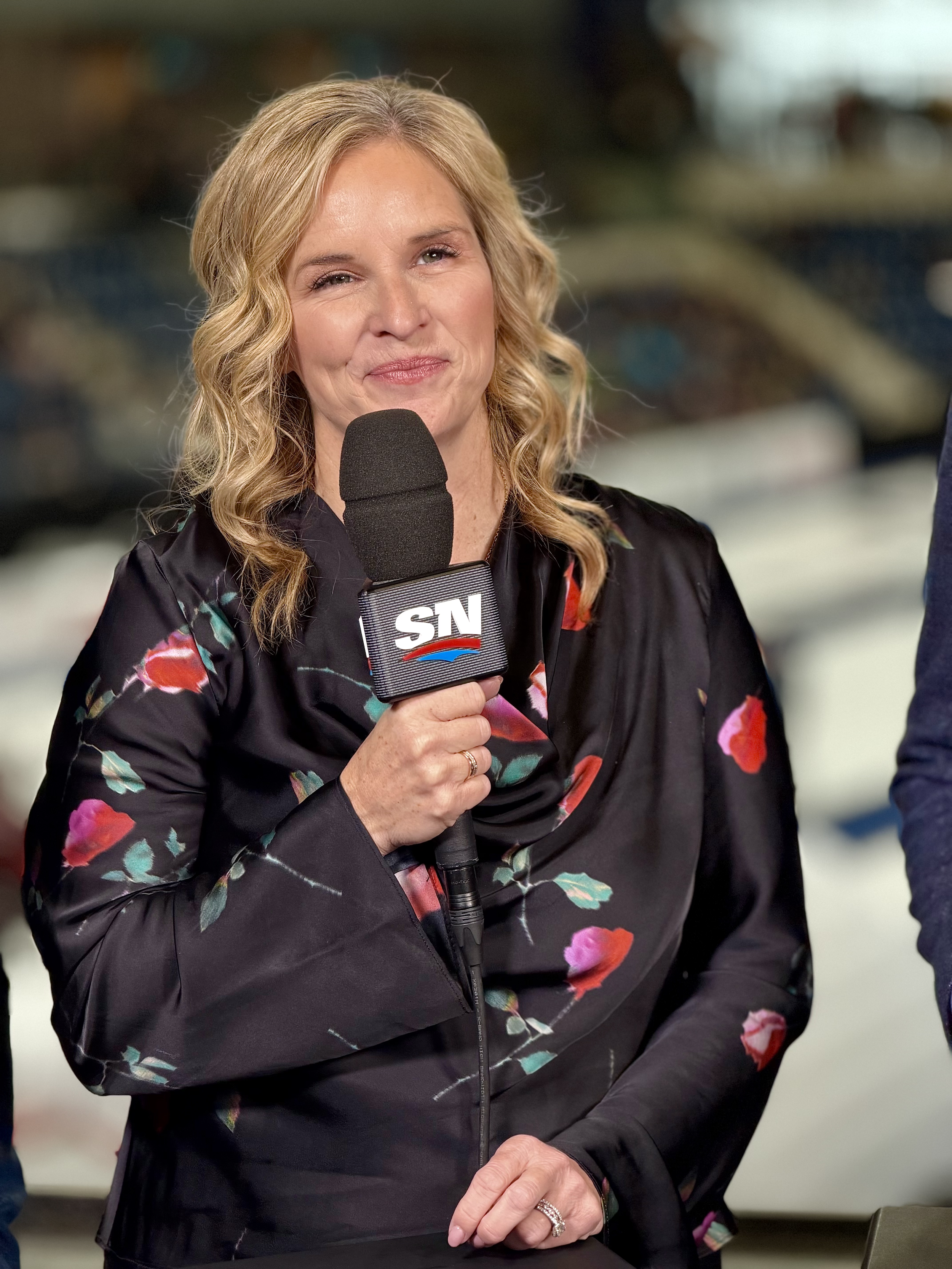 Jennifer Jones holding a microphone labeled 'SN,' wearing a black floral jacket, standing in a stadium or arena.