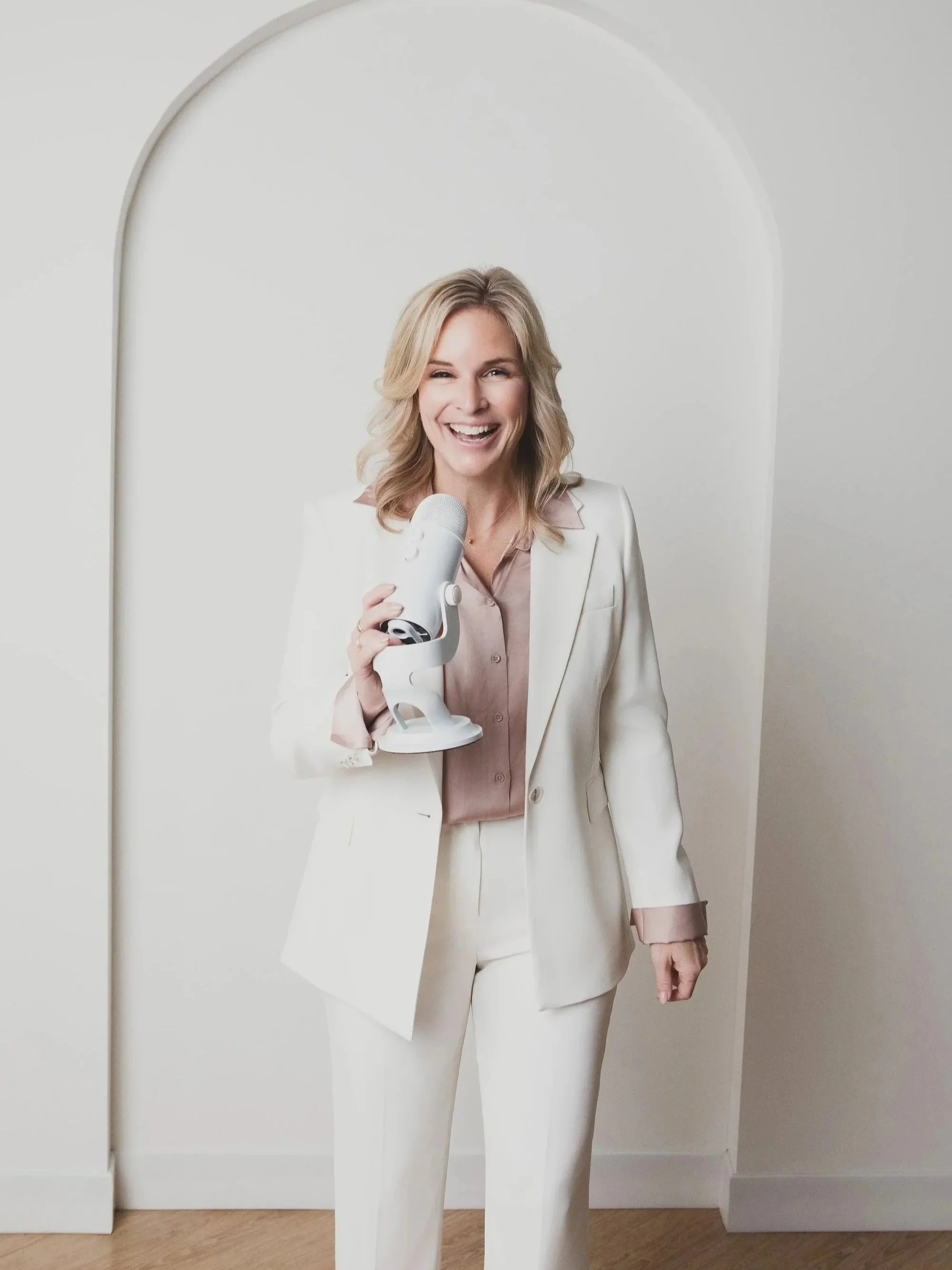 A woman with blonde hair, dressed in a cream-colored suit, smiling and holding a white microphone in a room with white walls and light wood flooring.