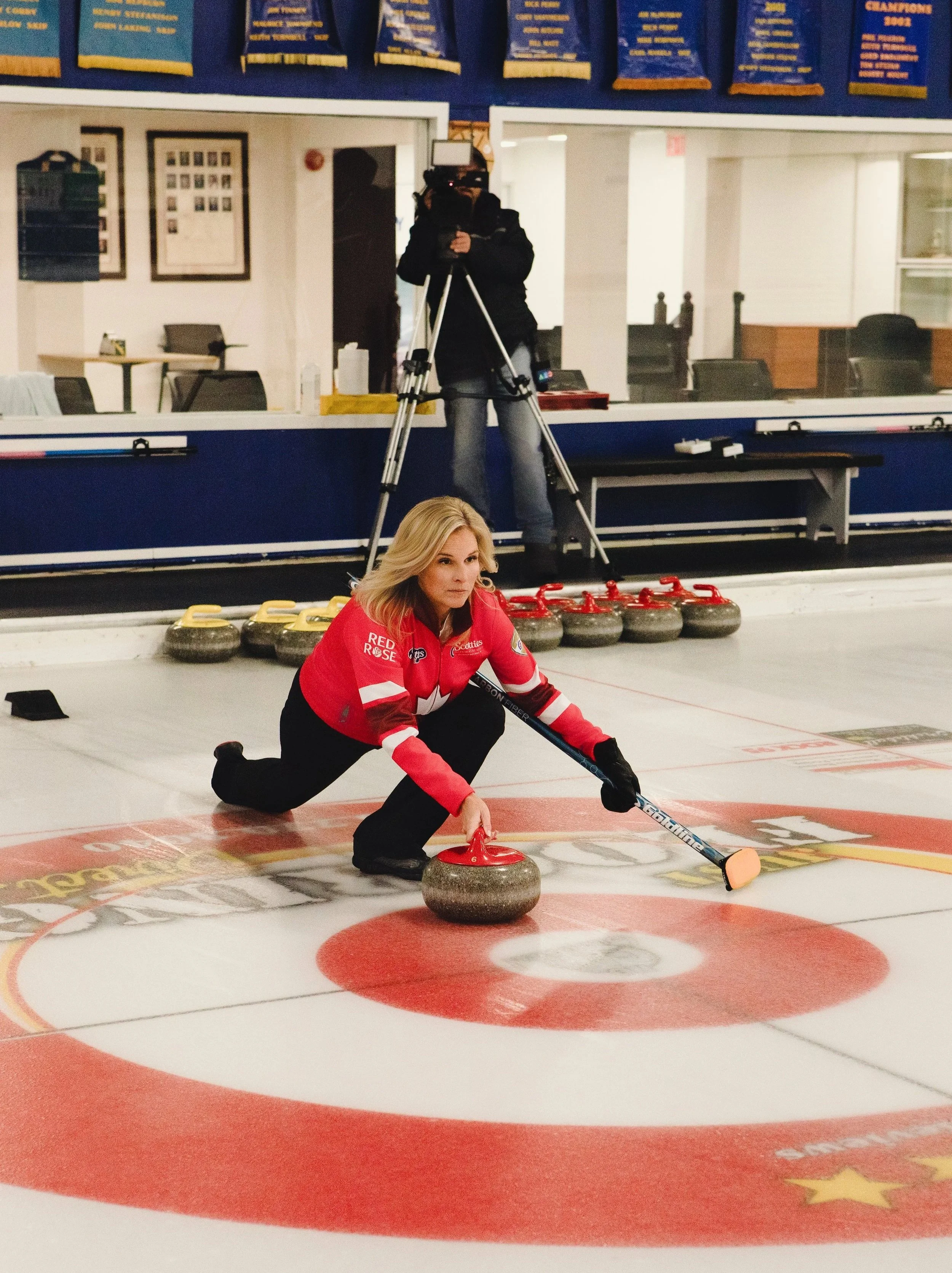 A woman in a red curling team jersey kneeling on an ice rink, about to release a curling stone towards the target area, with a cameraman filming her in the background.