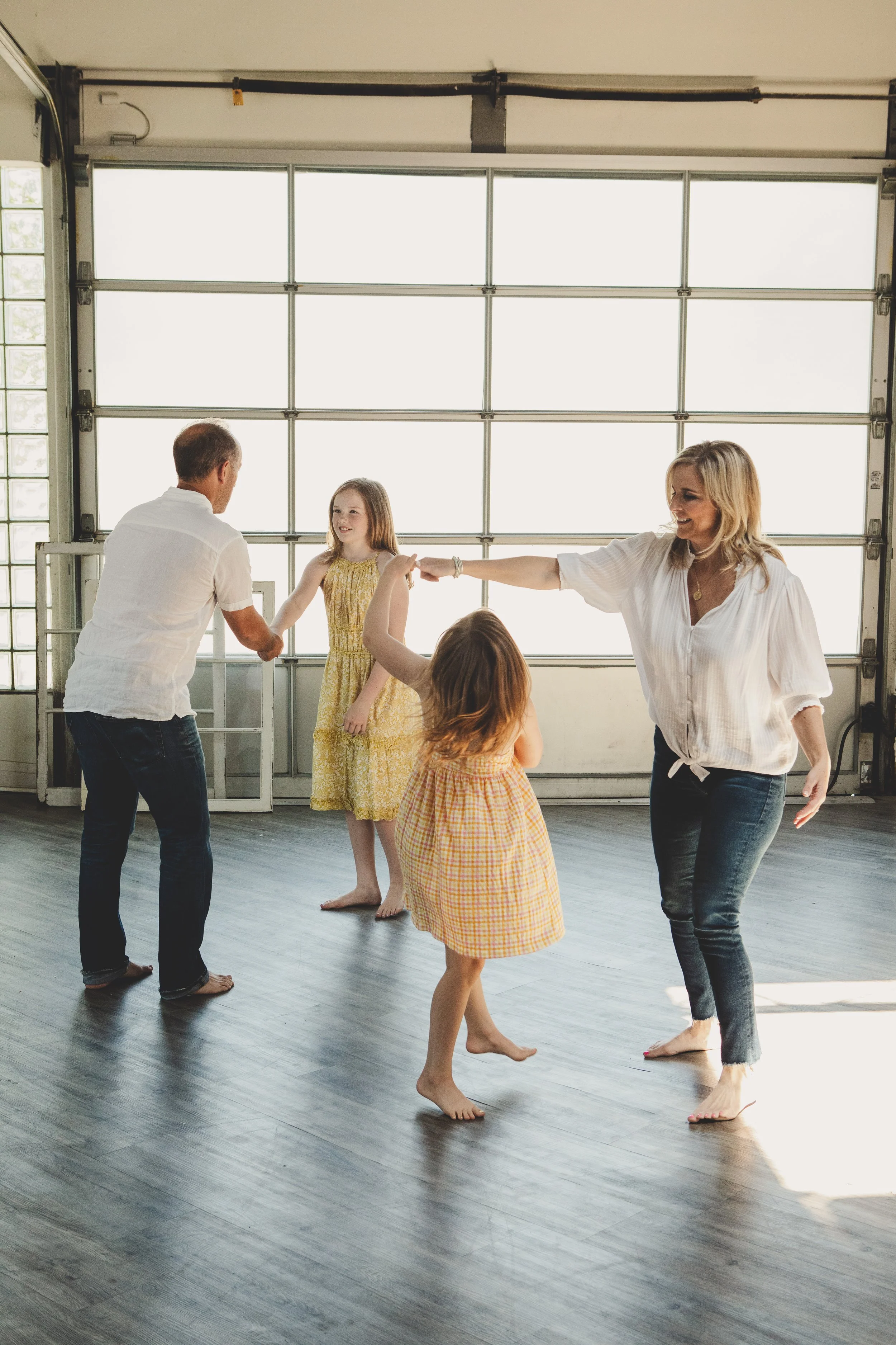 A family dancing and playing inside a garage with a large door, with children and a woman in the foreground and a young girl in a yellow dress standing in the background.