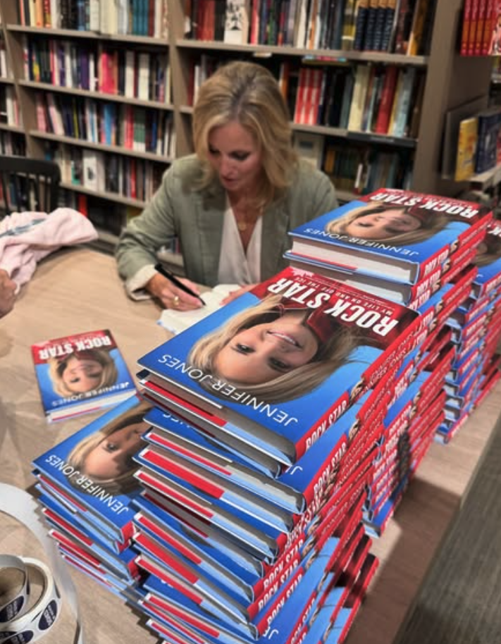 A woman signing copies of a book titled 'ROCK STAR' by Jennifer Joplin, with stacks of the same book on a table in a bookstore setting.