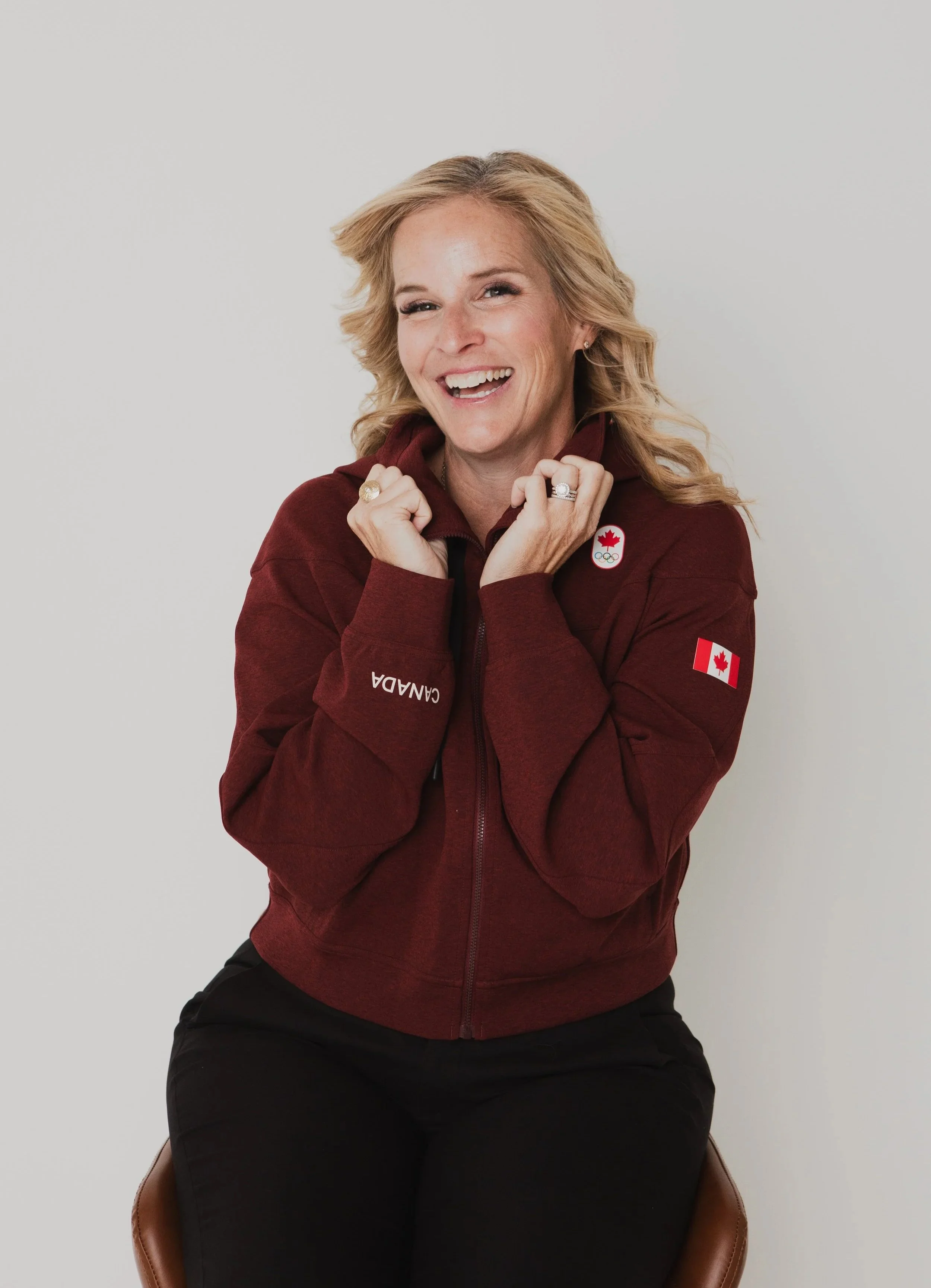 A woman with blonde curly hair smiling, wearing a maroon Canadian Olympic jacket with patches, sitting on a brown chair against a plain white background.