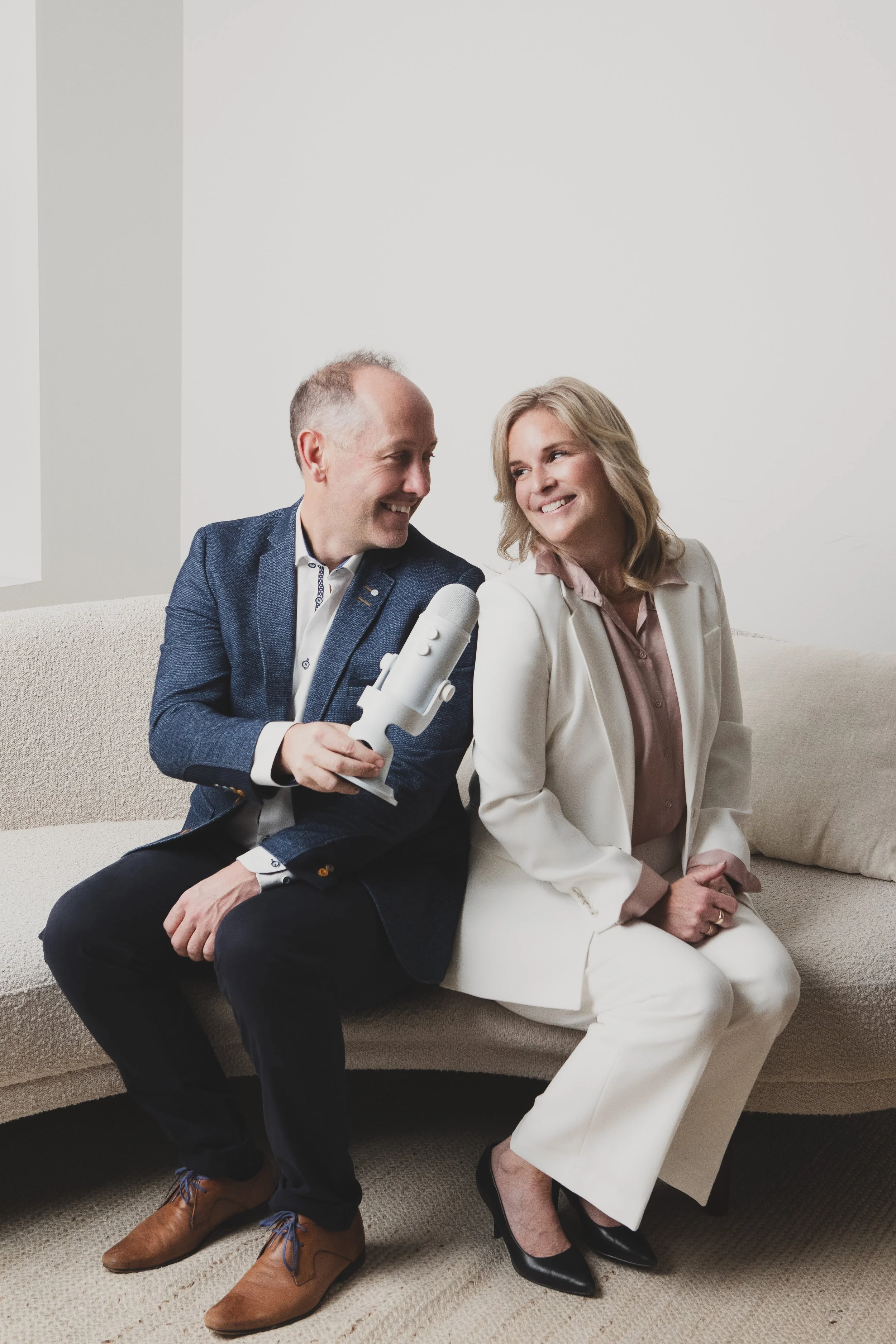 A man and woman sitting on a beige sofa, smiling at each other. The man is holding a white microphone and appears to be interviewing the woman, who is dressed in a white suit with a pink blouse. The room has white walls and a bright, minimalist setting.