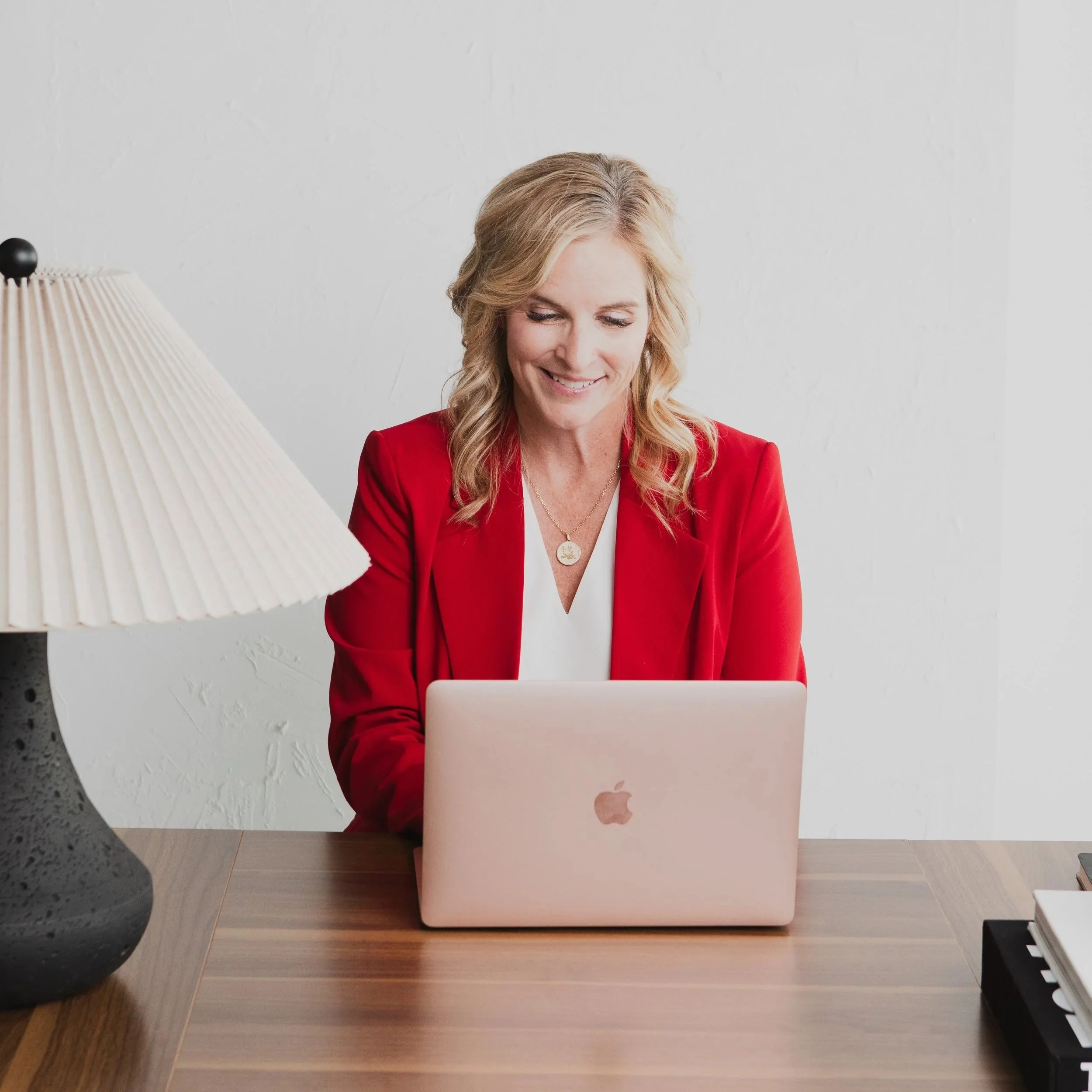 A woman with blonde hair wearing a red blazer and white top, sitting at a wooden desk, looking at her closed silver laptop, smiling.