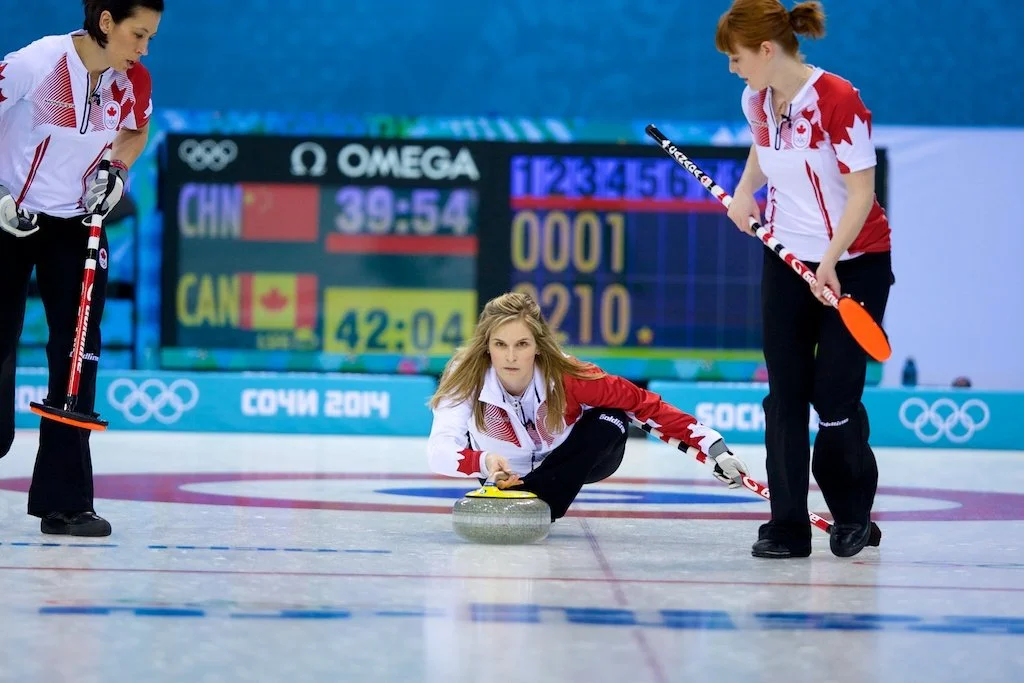 Olympic curling match with three female athletes, one kneeling and delivering a curling stone, another standing preparing to sweep, and the third observing, with scoreboard in background showing China and Canada scores, during the 2014 Sochi Winter Olympics.