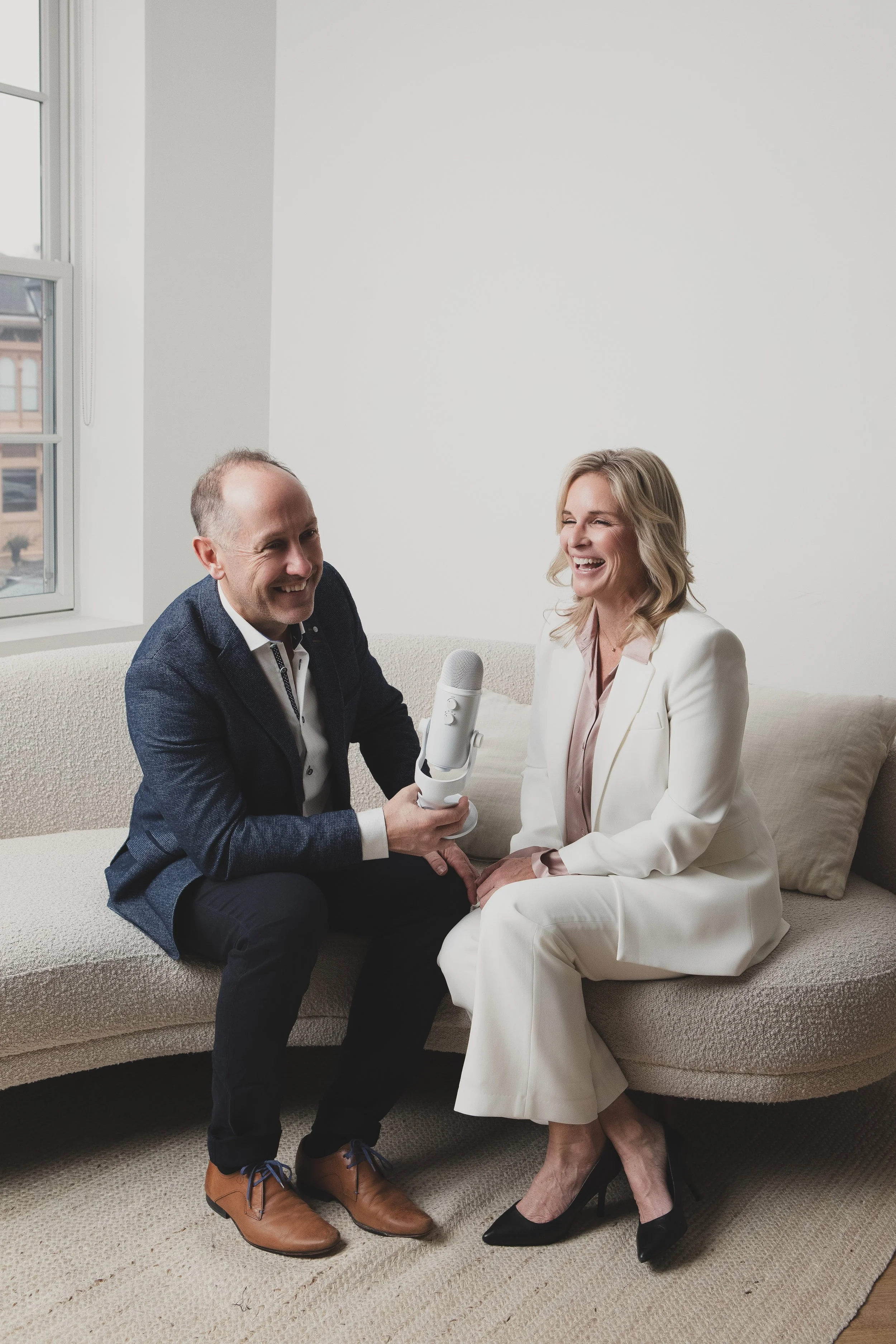 A man and woman sitting on a beige couch, smiling and laughing during a podcast or interview, with a microphone between them in a bright room with a window.