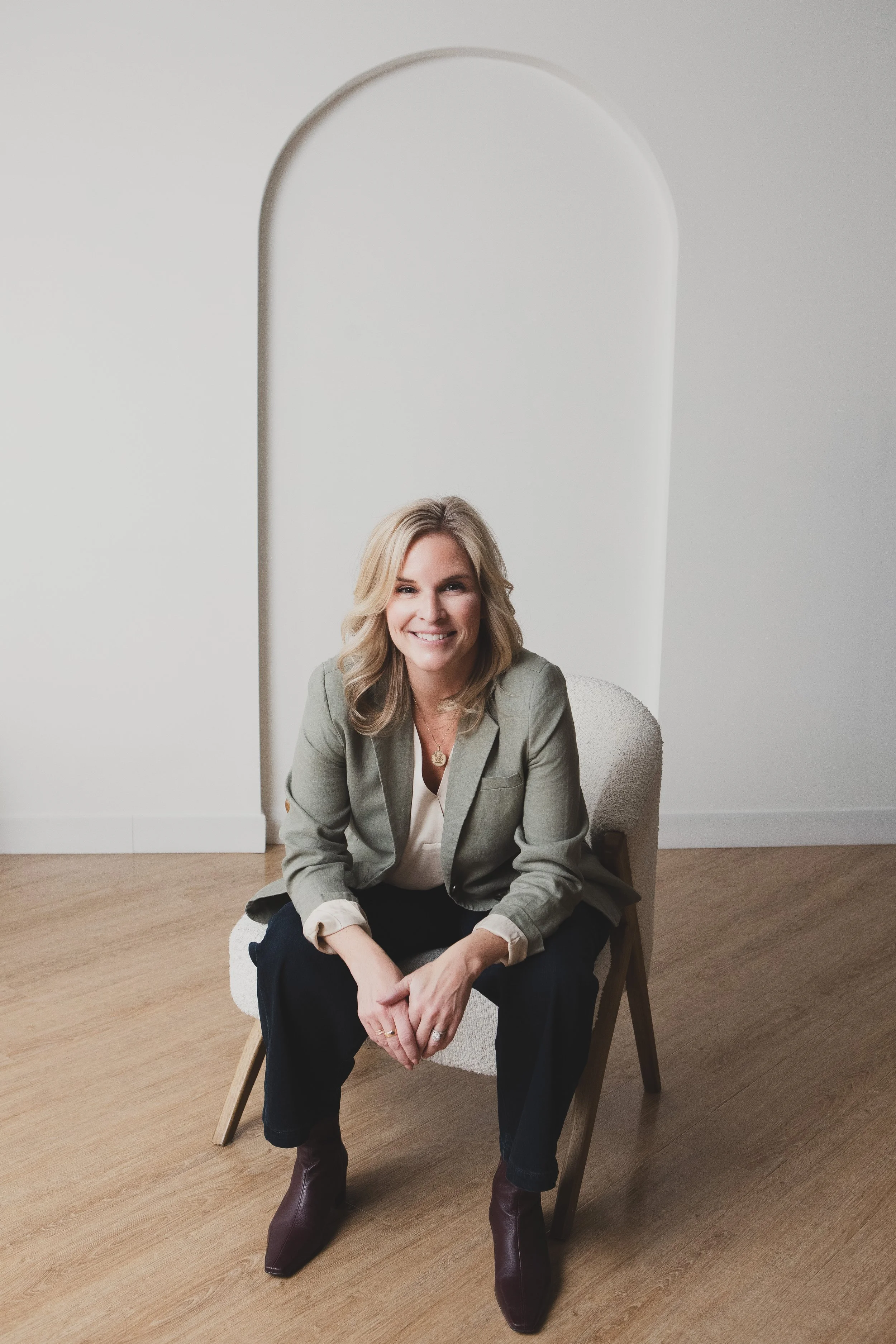 A woman with blonde hair, wearing a light gray blazer, white shirt, dark pants, and brown boots, sitting on a chair in a room with light-colored walls and wooden flooring, smiling at the camera.