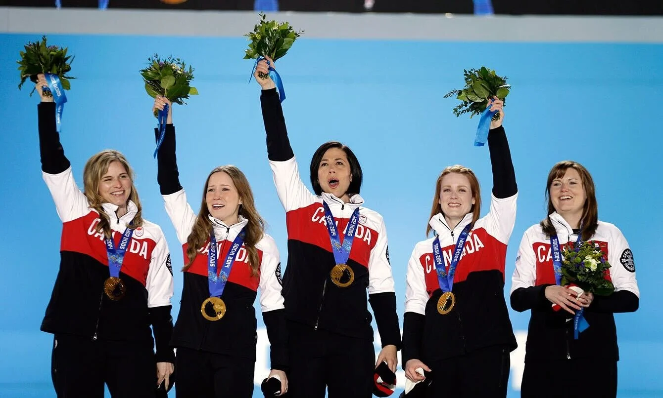 Five female athletes in Team Canada uniforms standing on a podium with medals, holding bouquets, celebrating victory at a sports event.