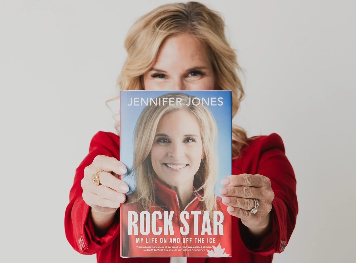 Woman with blonde hair in a red jacket holds a book titled 'Rock Star' with a photo of herself, partially obscuring her face, against a plain gray background.