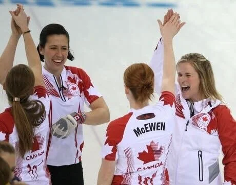 Canadian female athletes celebrating with high-fives, wearing red and white uniforms with maple leaf insignia.