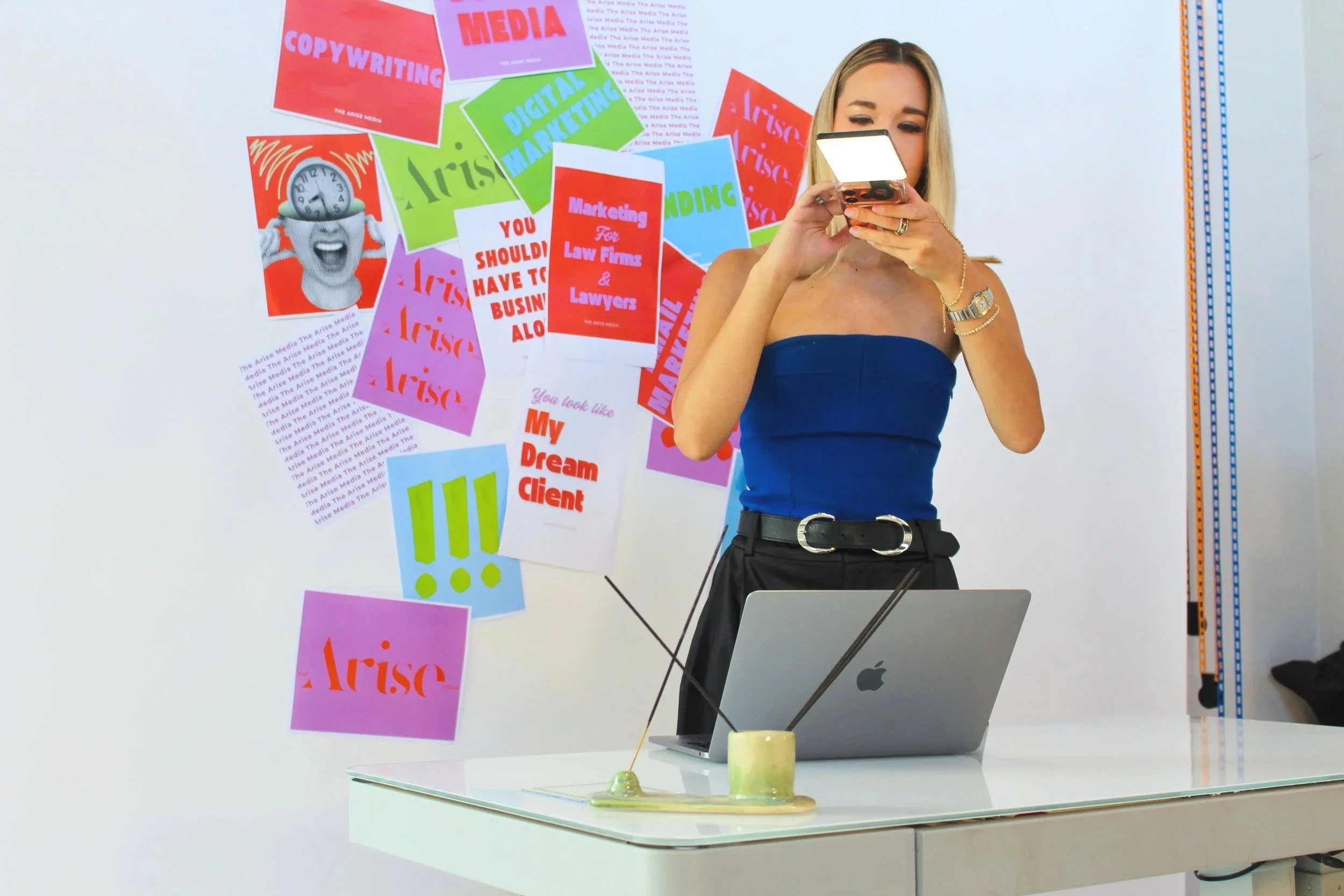 A woman in a blue strapless top and black pants is standing at a white table, looking at her phone. Behind her, a wall is decorated with colorful posters and flyers related to media, copywriting, digital marketing, and law firms. There is an open laptop and a cup of tea or coffee on the table.