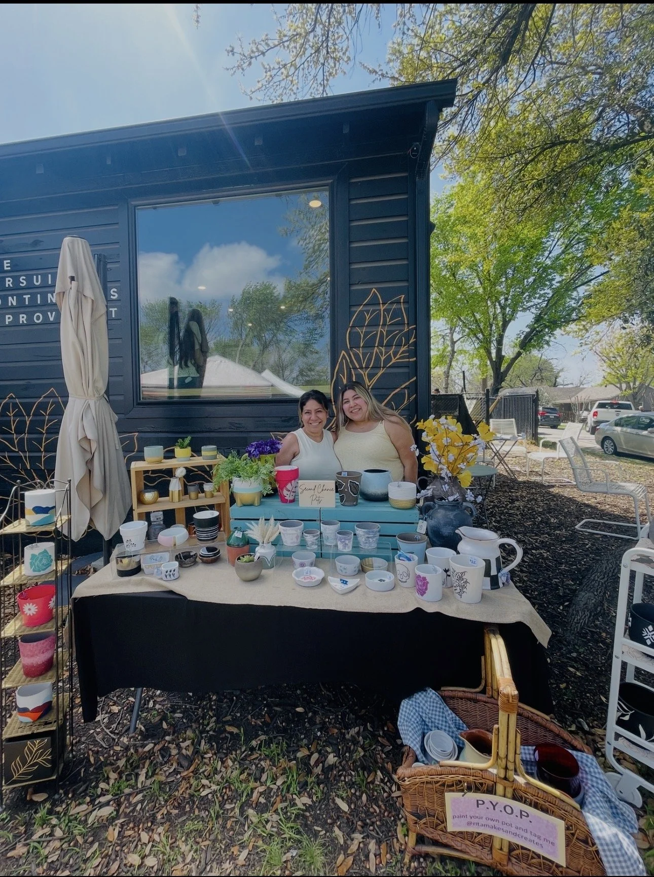 Two women standing behind a table of plant pots at an outdoor craft fair, with trees and parked cars in the background.