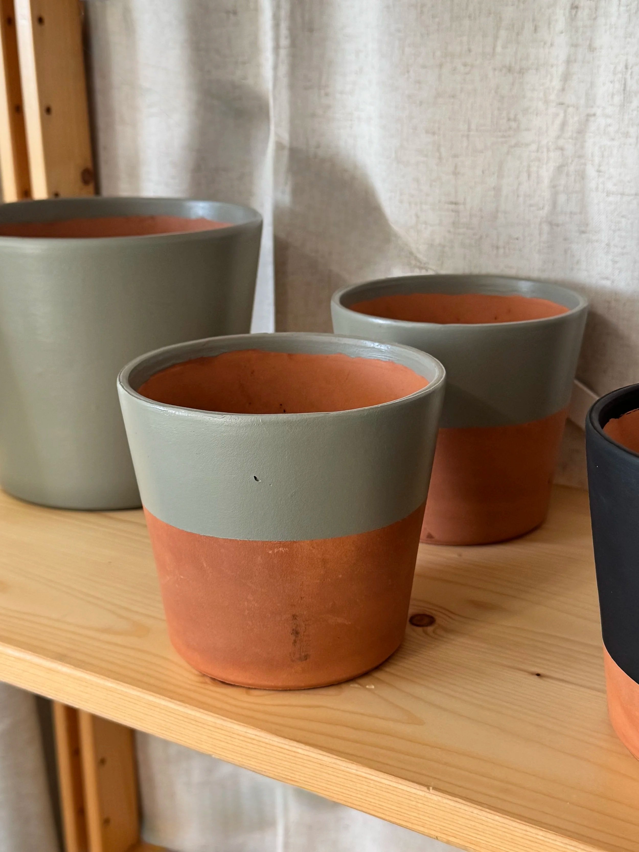 Set of four plant pots in muted green, terracotta, and black colors displayed on a light wooden shelf against a beige curtain.
