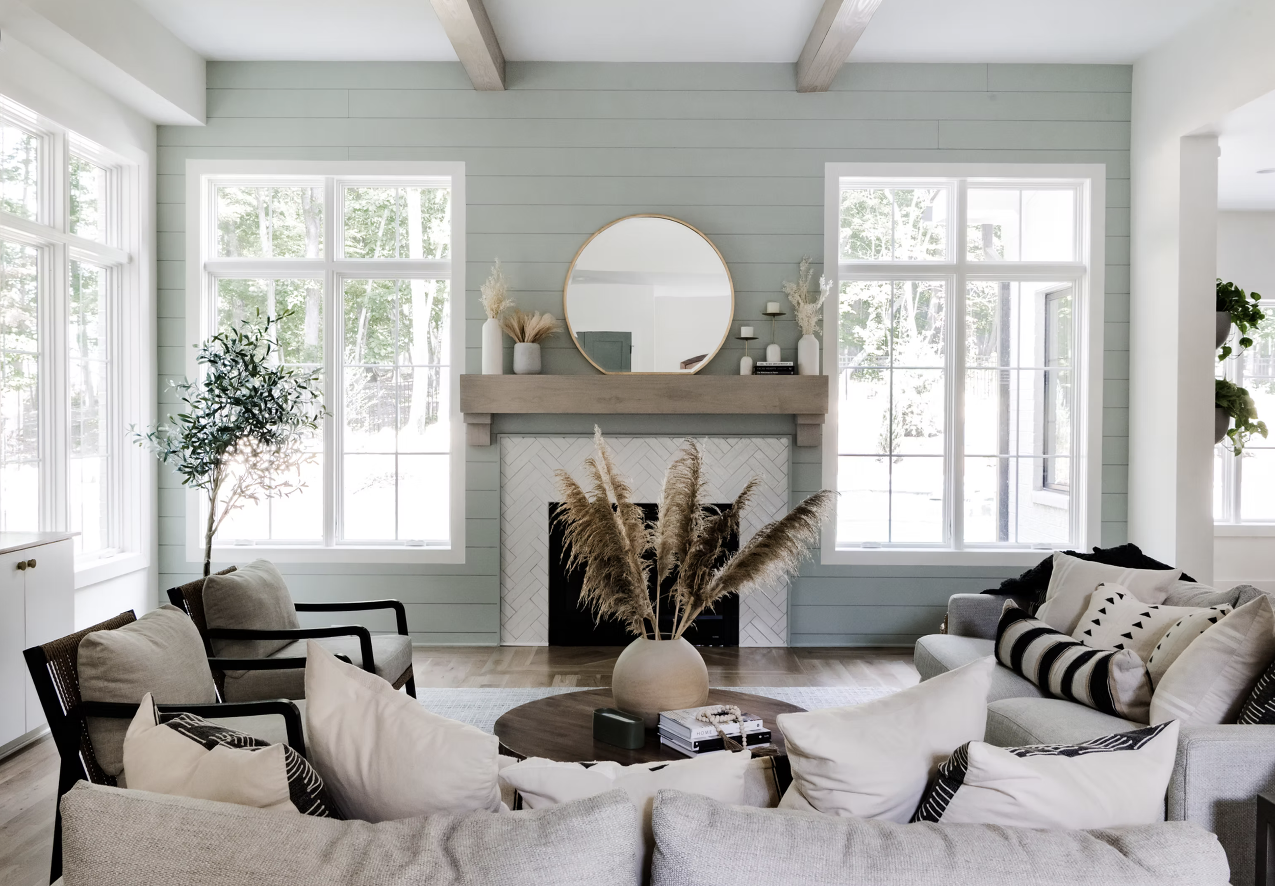Living room with light green shiplap walls, large windows, a fireplace with a round mirror above, and beige sofas with black-and-white pillows. Decor includes vases with dried plants and candles, a potted plant on the right, and a large white ceramic vase with pampas grass on a round wooden table.