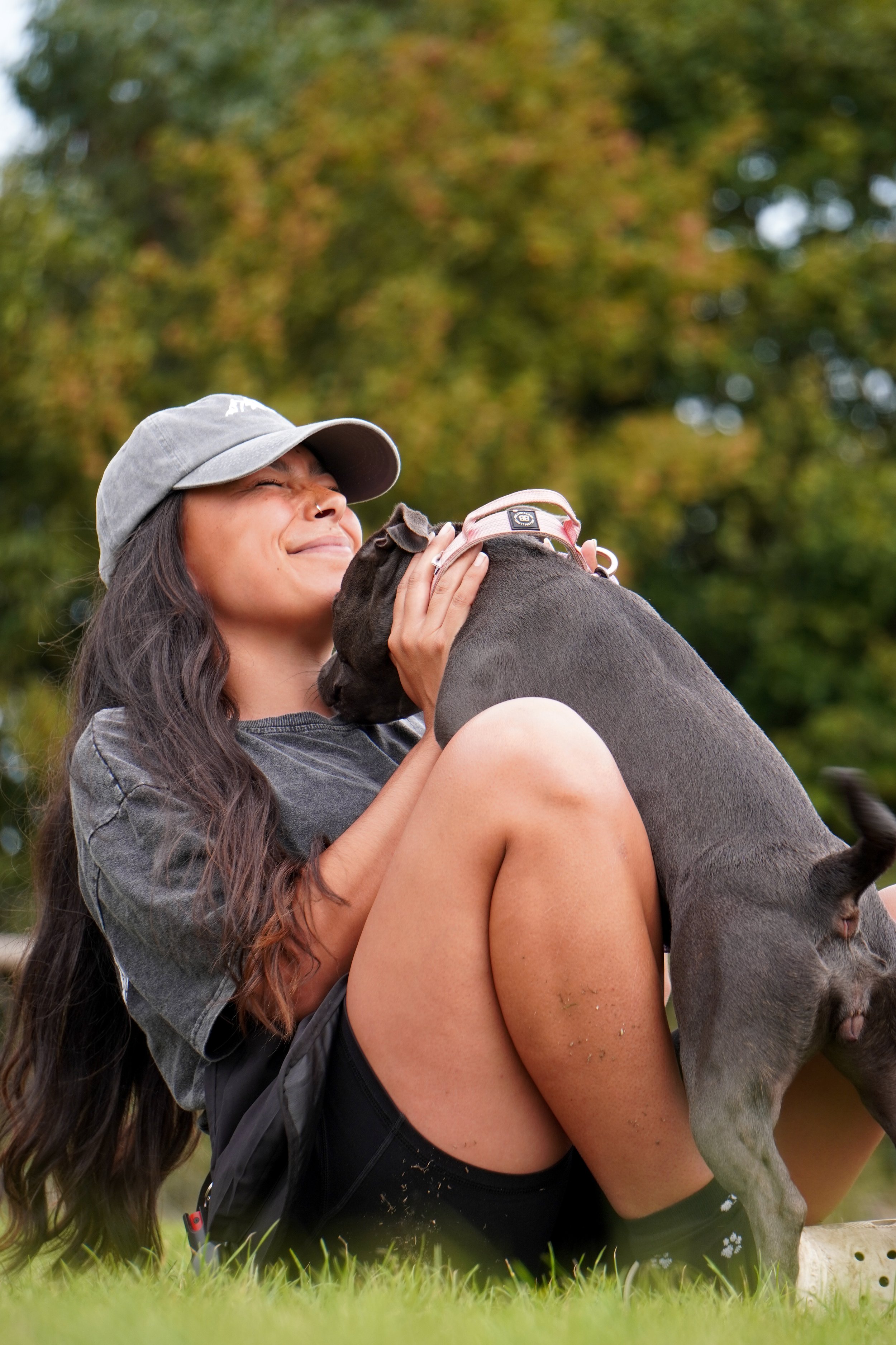 Lead & Ramble. A woman outdoors smiling and hugging a small grey dog with a pink collar, sitting on grass with trees in the background.