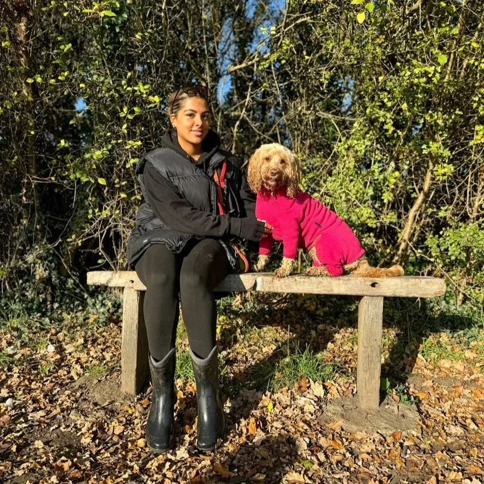 A woman in a black jacket and black rain boots sitting on a wooden bench outdoors with a small golden doodle dog dressed in a pink sweater and pants, surrounded by trees and fallen leaves.