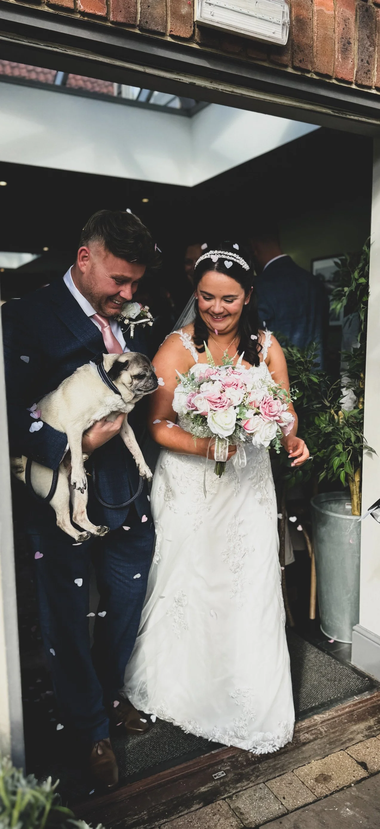 A newlywed couple celebrates their wedding with a small, light-colored dog, surrounded by flower petals and guests, indoors near the entrance.
