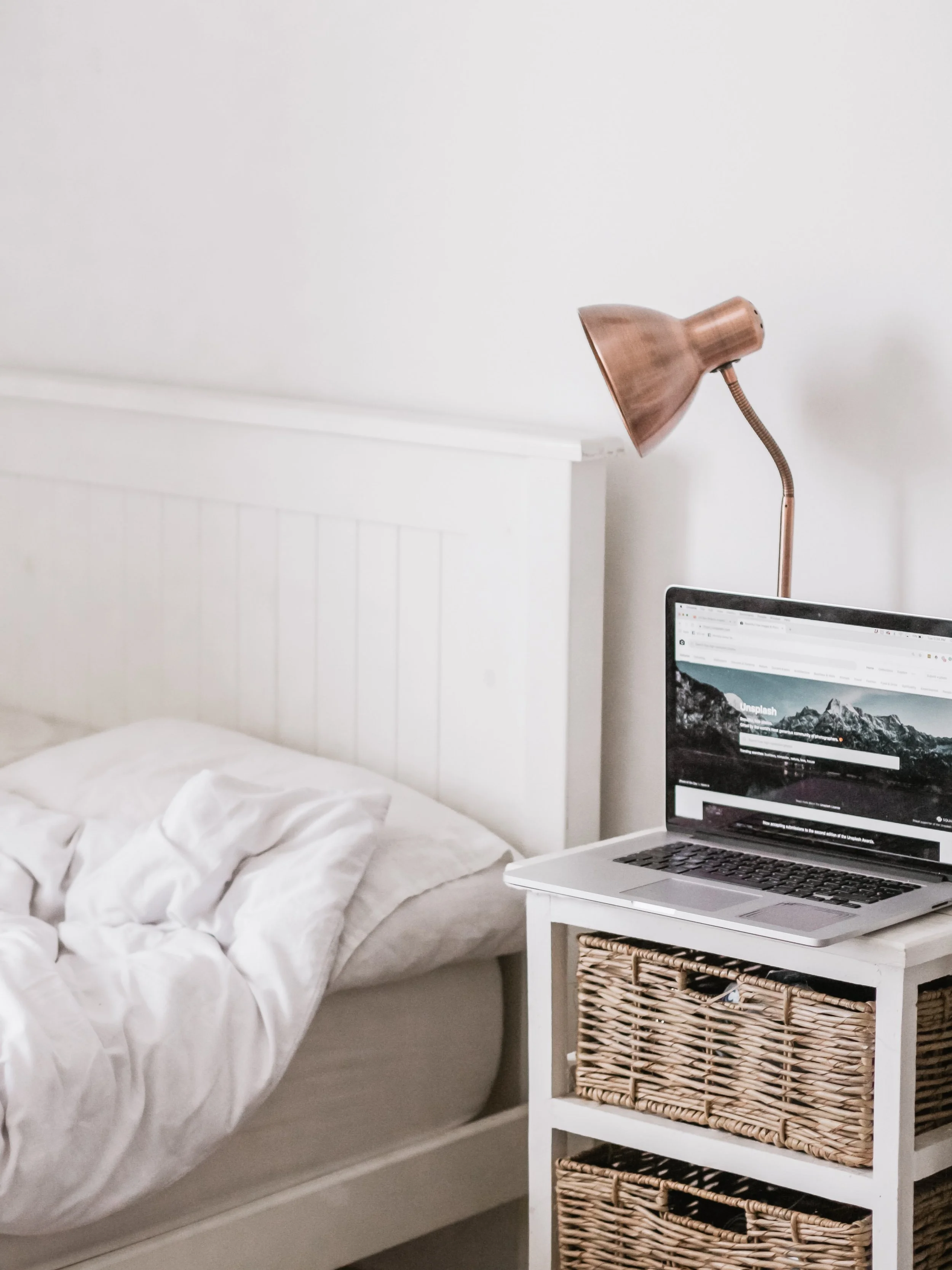 A bedroom bedside table with a laptop, a copper desk lamp, and a bed with white bedding against a white wall.