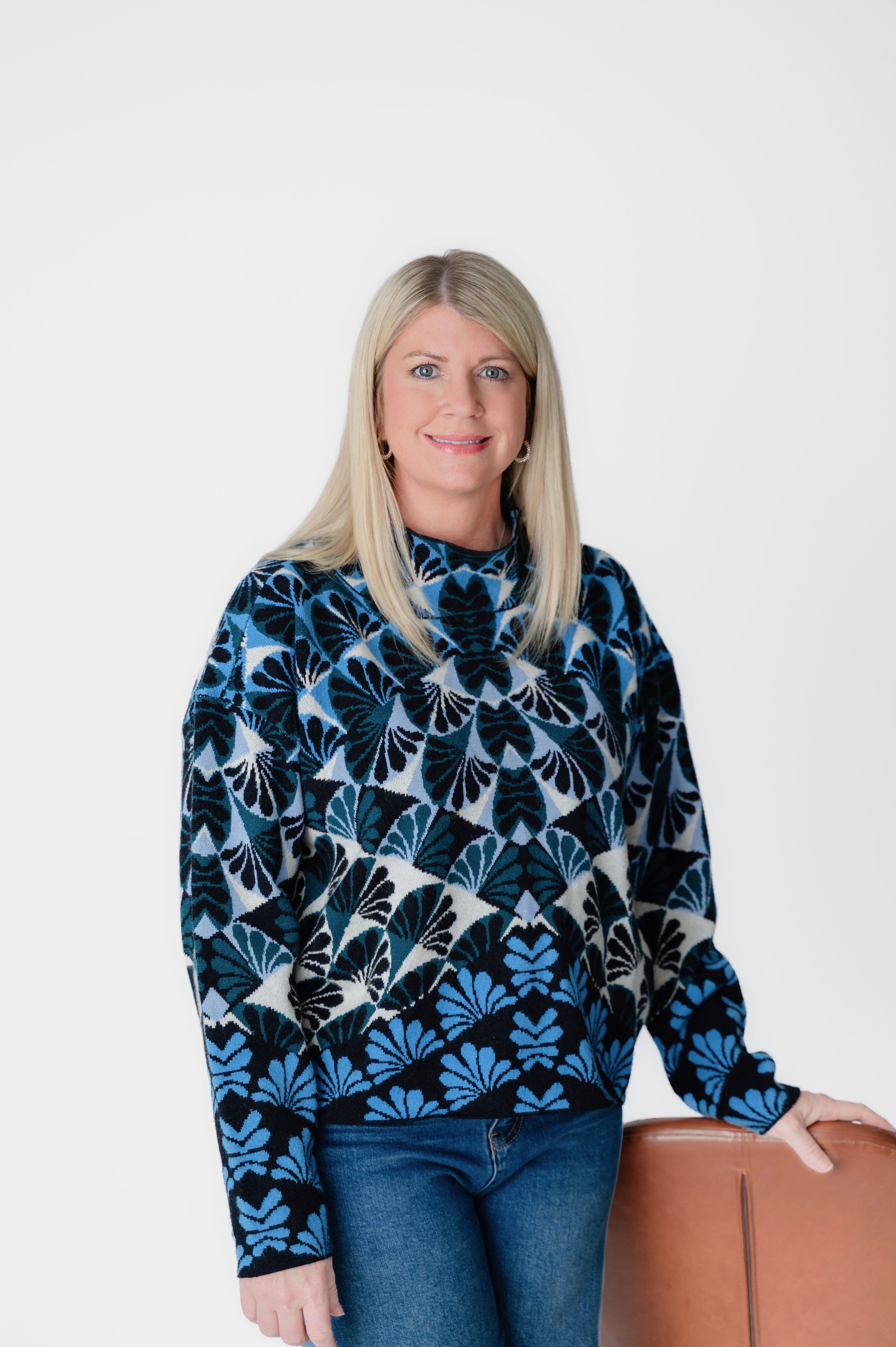 A blonde woman in a blue and black patterned sweater and jeans standing next to a brown chair against a white background.