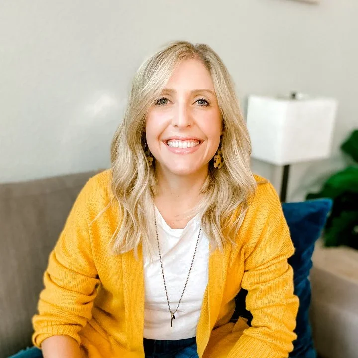 A woman with blonde hair smiling, wearing a yellow cardigan, sitting on a couch in a cozy room with a lamp and pillows.