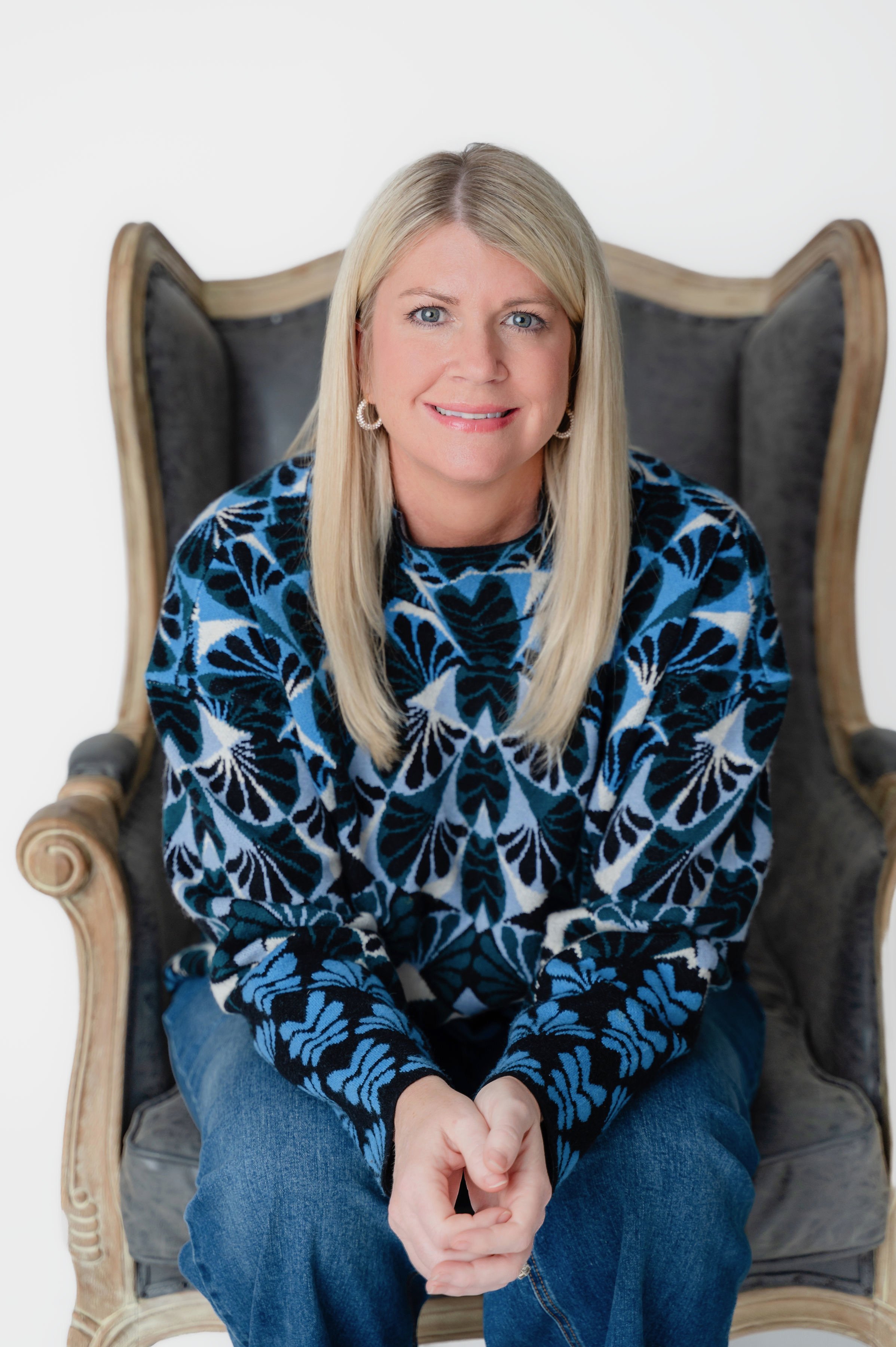 A woman with blonde hair sitting on a vintage high-back chair with dark upholstery, wearing a patterned blue and black sweater and blue jeans, smiling at the camera.