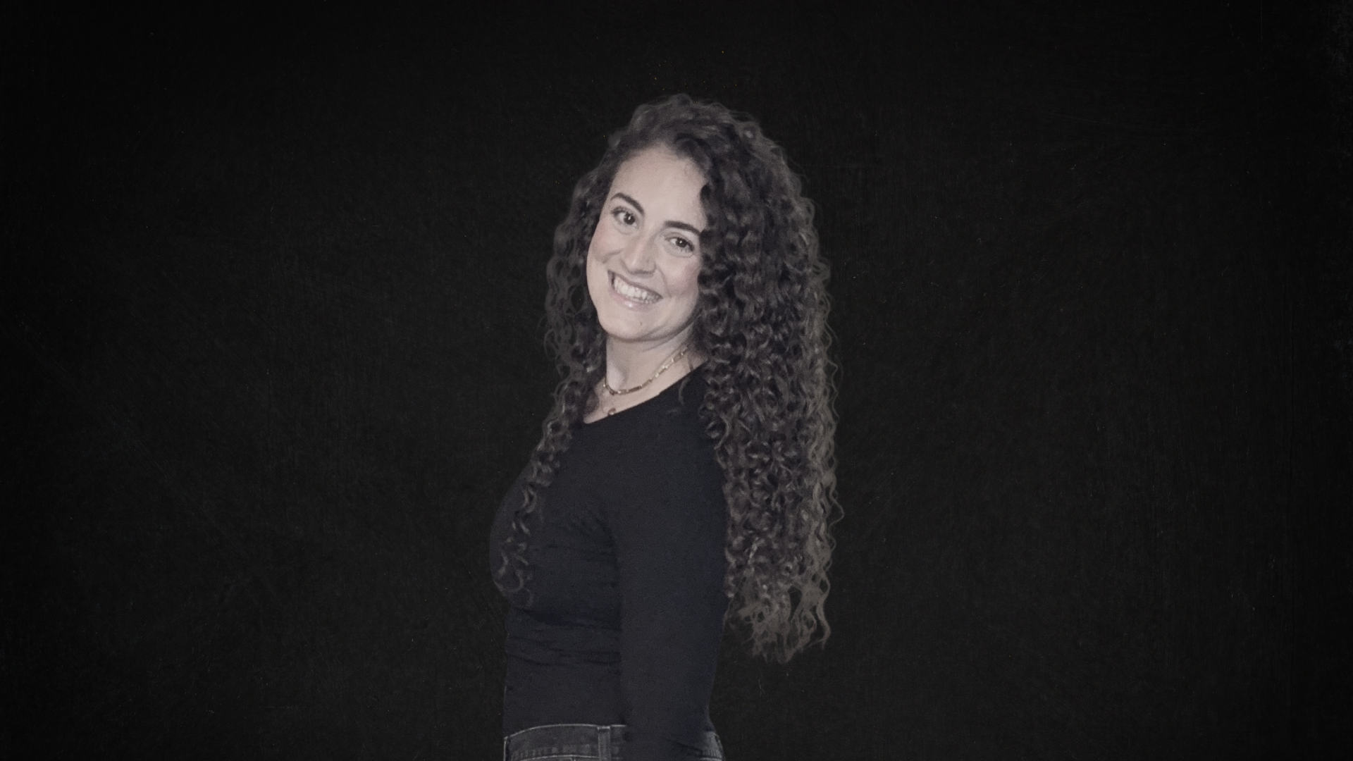 A woman with long, curly brown hair smiling and posing against a dark background, wearing a black top and a necklace.