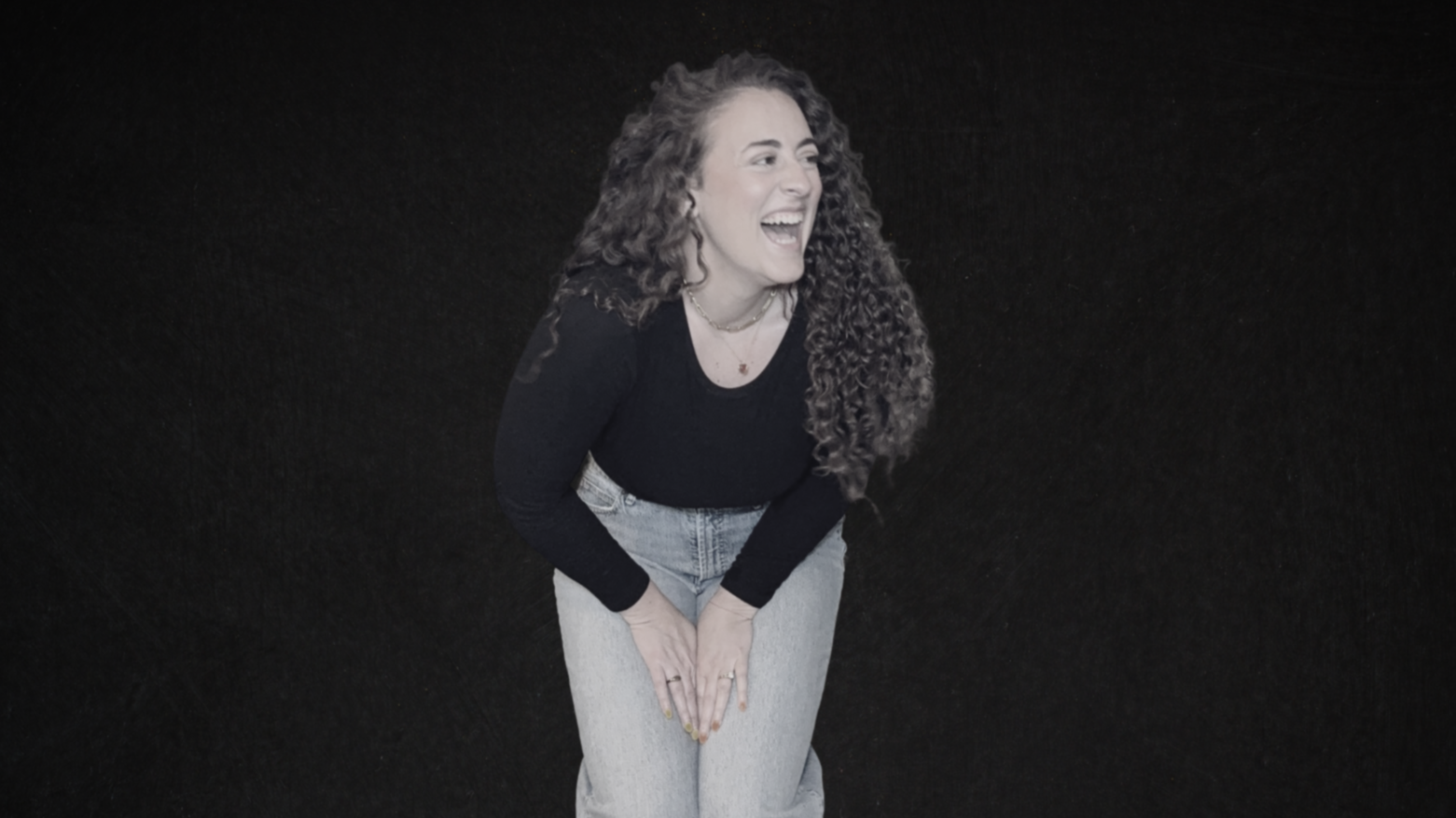 A woman with curly hair laughing and leaning forward against a dark background.