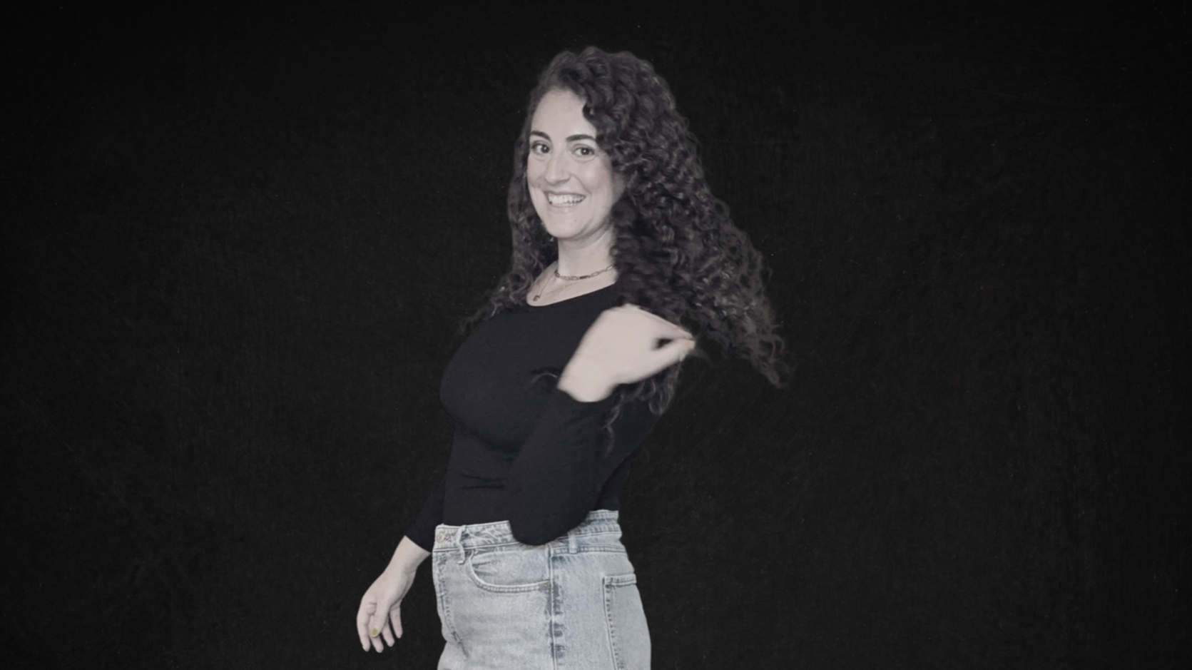A young woman with long, curly dark hair, smiling, wearing a black long-sleeve shirt and light jeans, standing against a dark background.