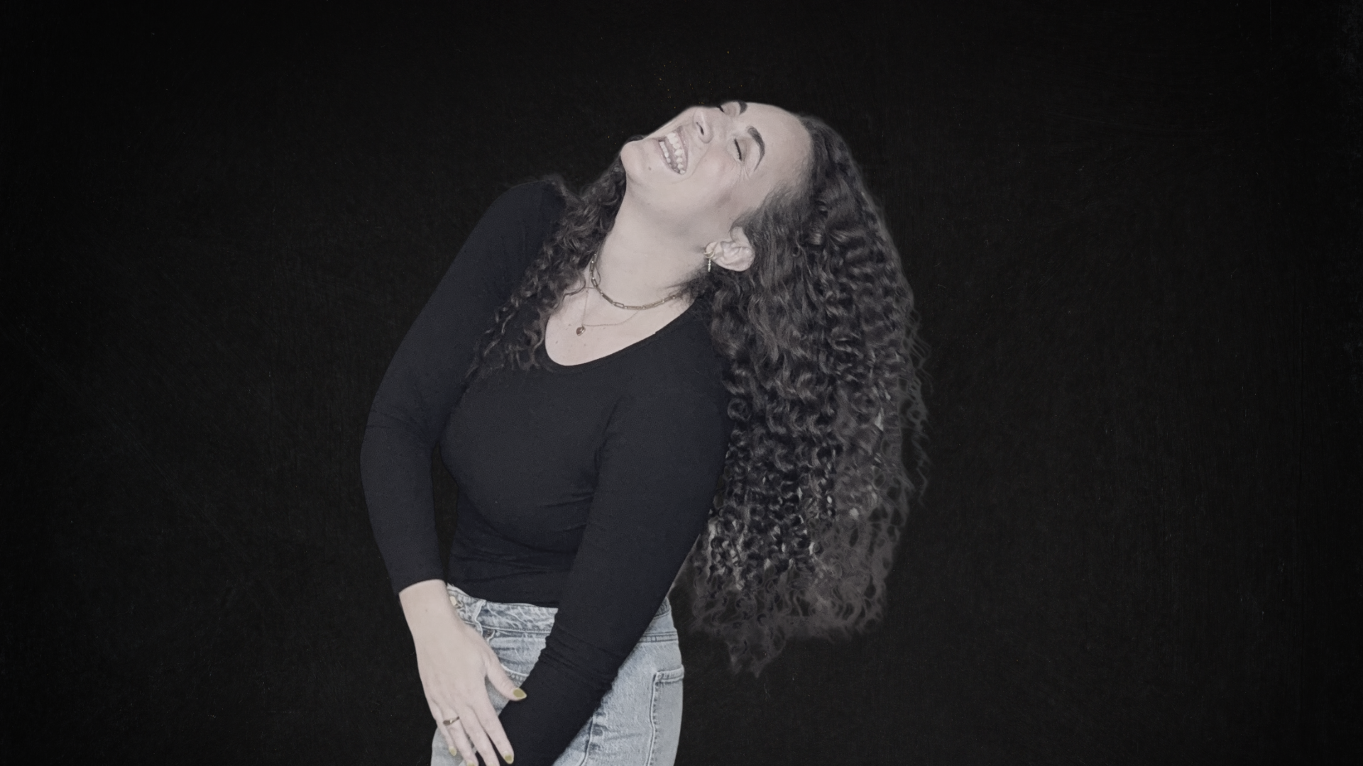 A woman with long, curly hair wearing a black long-sleeved top and light-colored jeans, smiling and tilting her head back against a black background.