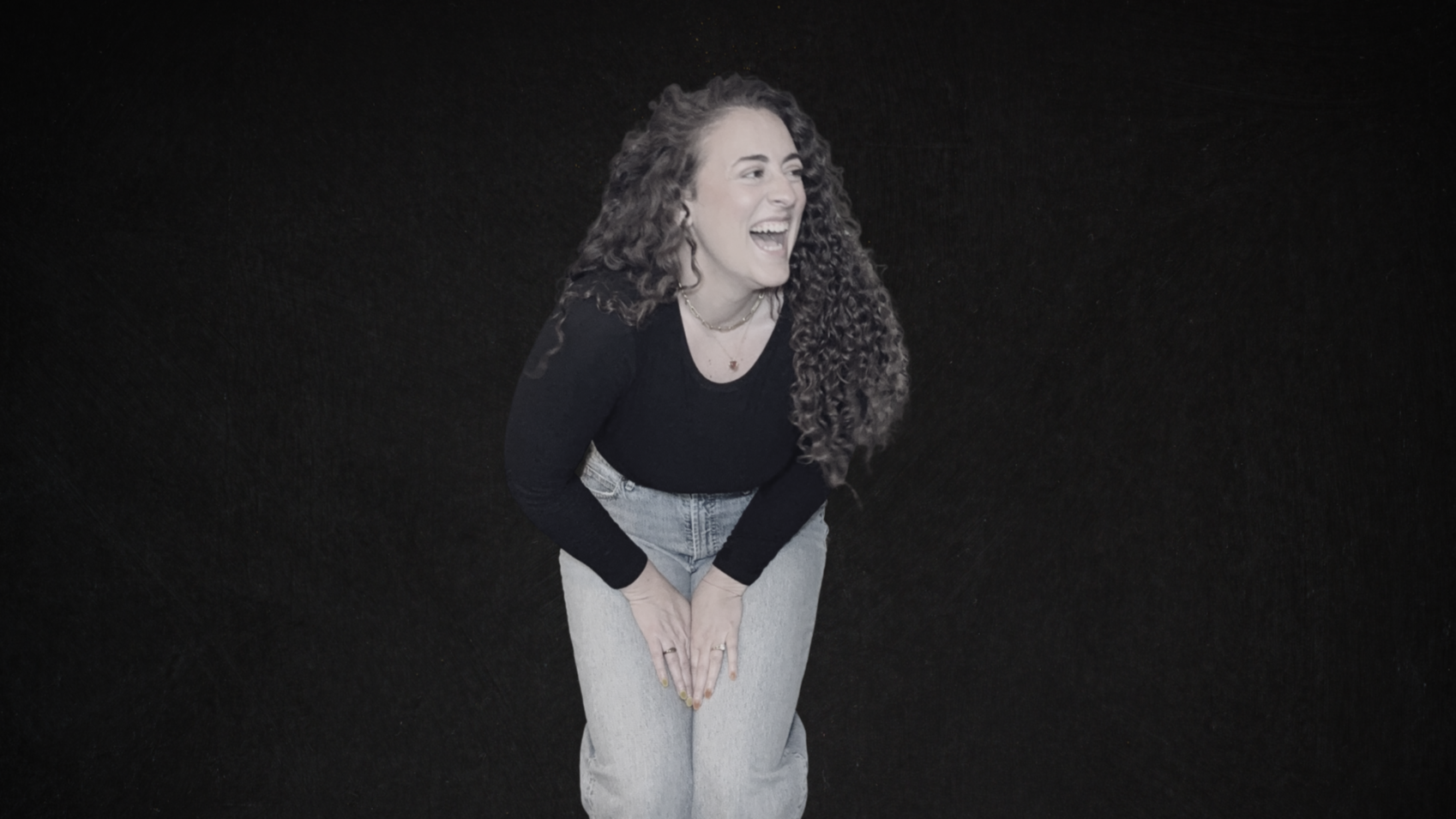 A young woman with long curly hair, wearing a black long-sleeve top and light jeans, is laughing and leaning forward against a black background.