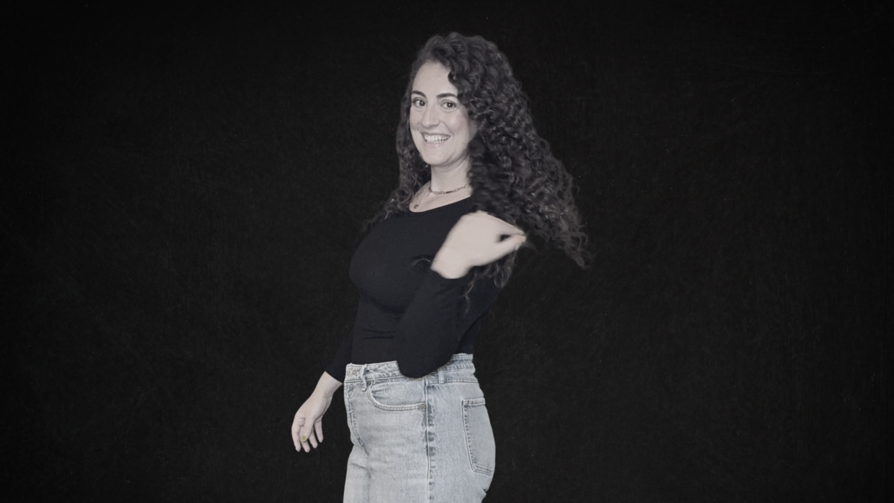 Young woman with curly hair smiling and looking at the camera, wearing a black top and light-wash jeans, standing against a black background.