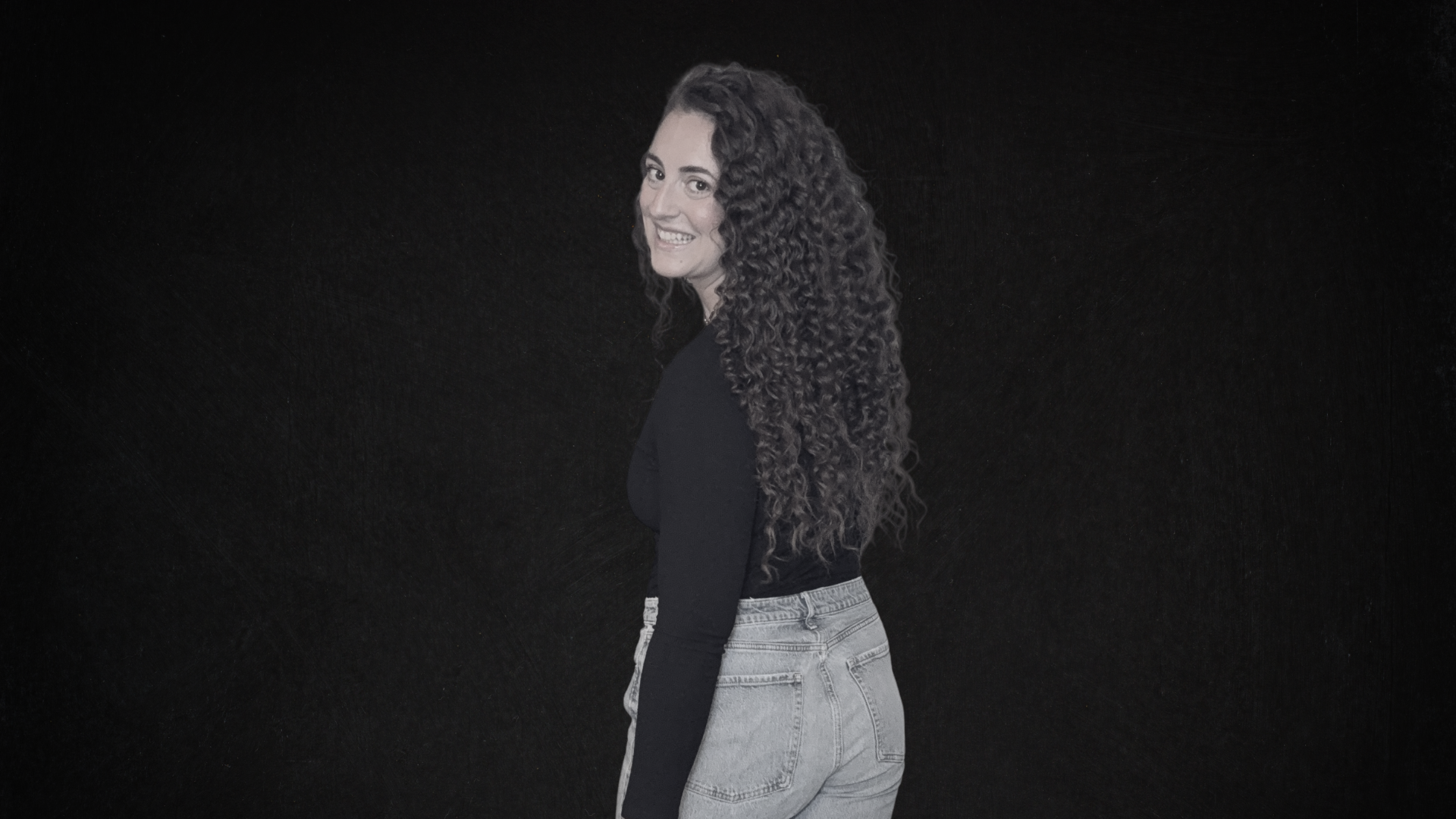 A woman with long, curly hair smiling while standing against a black background.