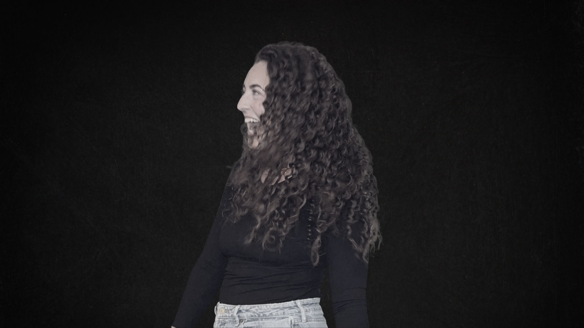 Side profile of a woman with long curly hair, smiling, against a dark background.