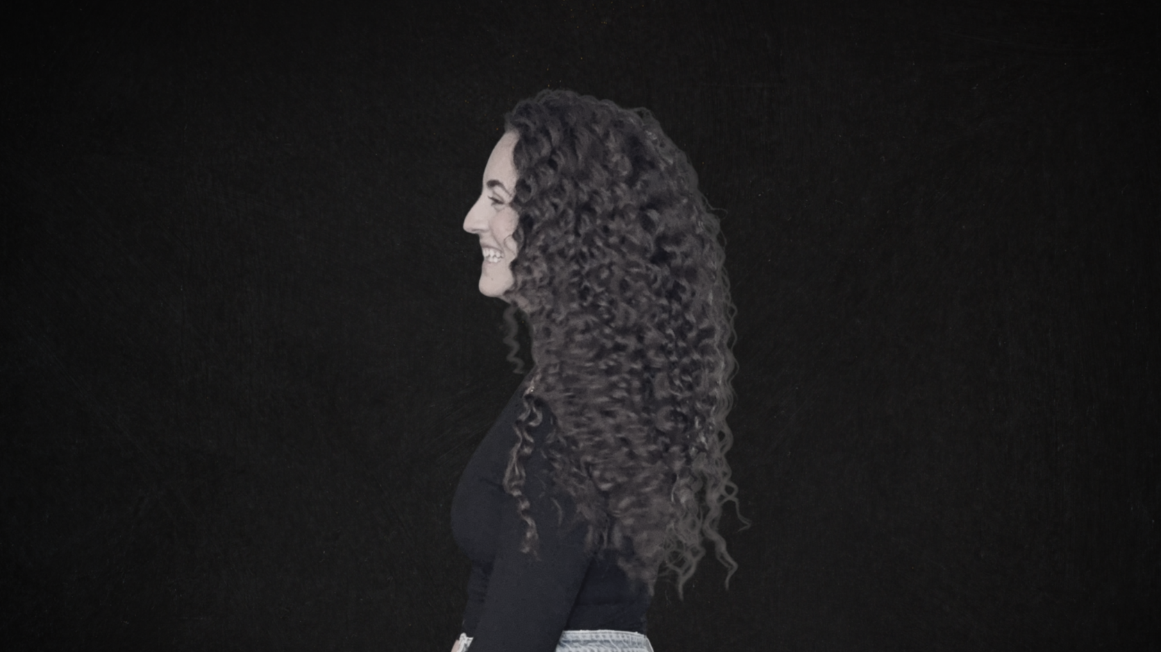 Profile of a smiling woman with long, curly hair against a black background.