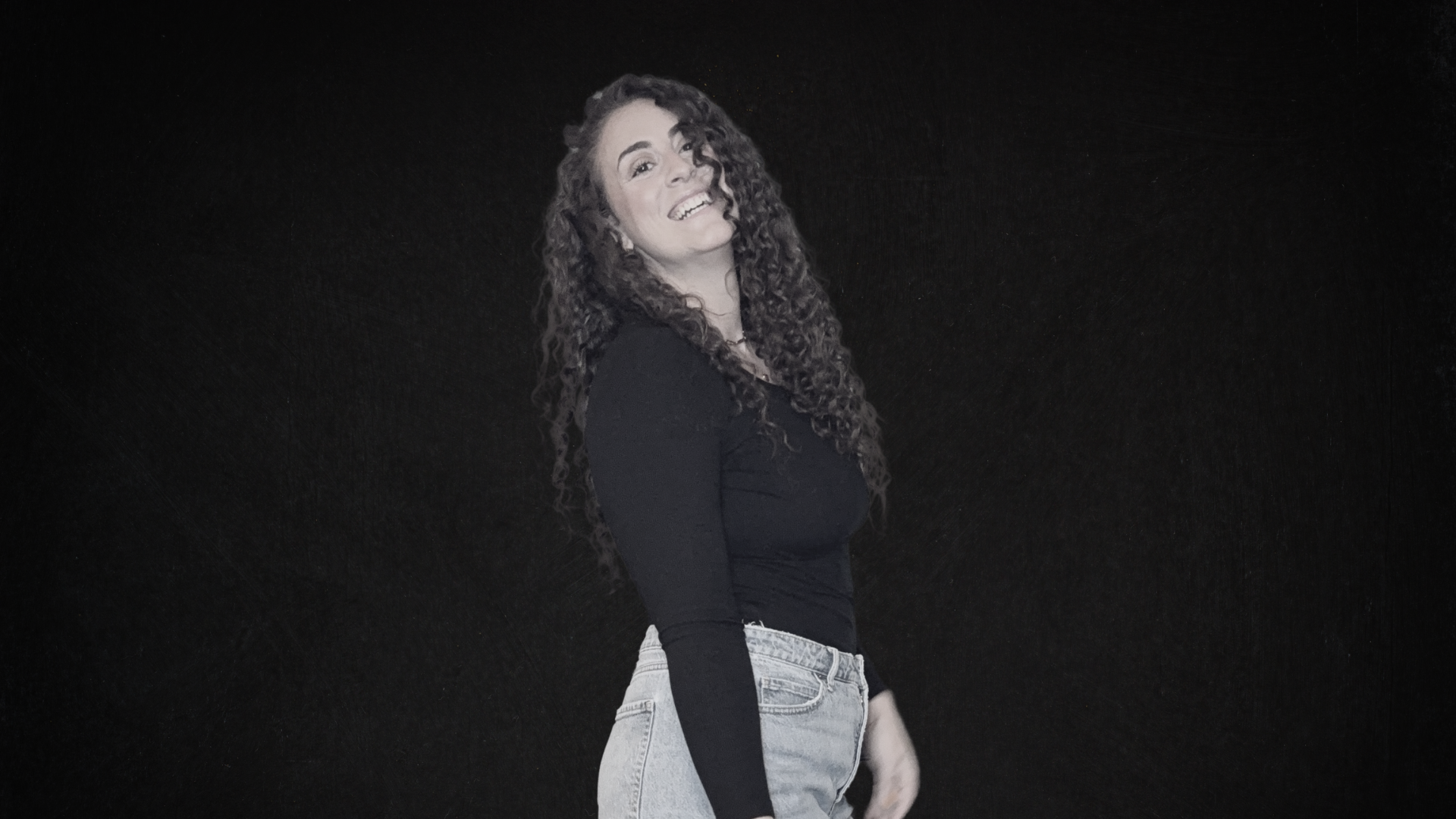 Young woman with long, curly hair smiling and looking at the camera, standing against a plain black background.